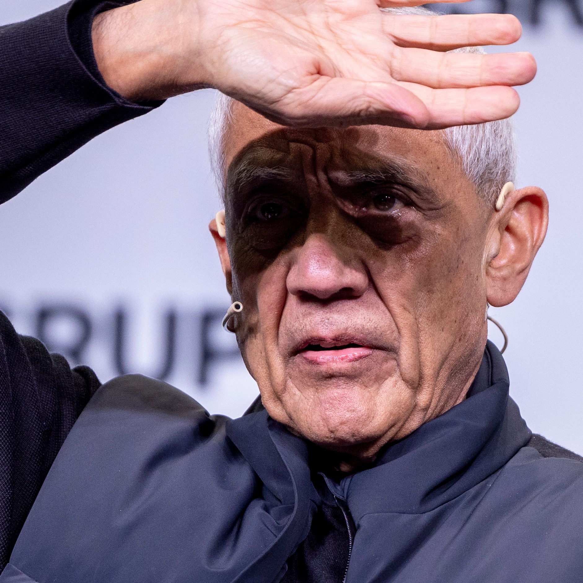 An elderly man with short gray hair holds his hand over his forehead. He is wearing a dark jacket and appears to be on stage, with a microphone in his ear.