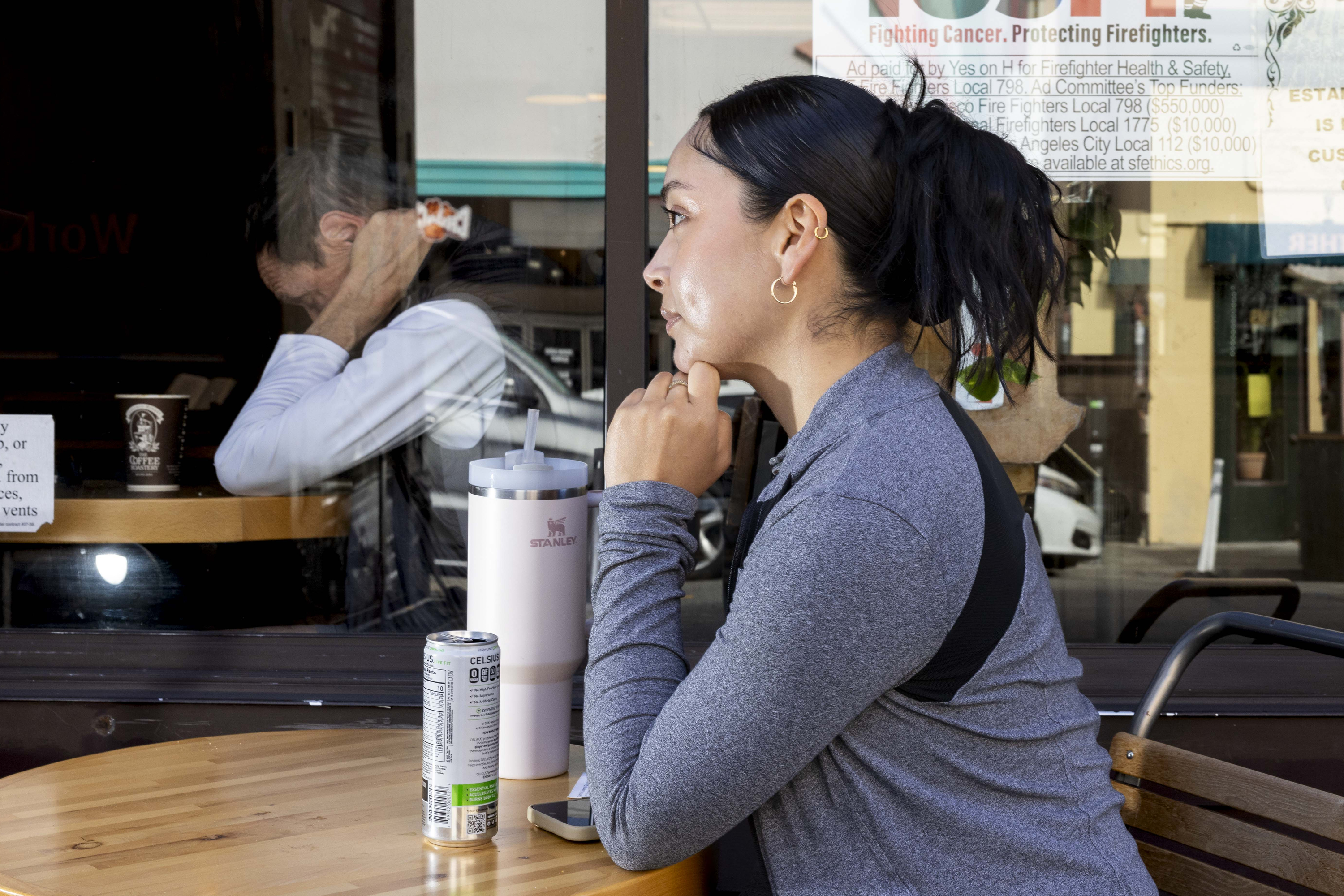 A woman with a ponytail sits at an outdoor cafe table with drinks. Behind her, a man is inside, holding his head in his hands, reflected in the window.