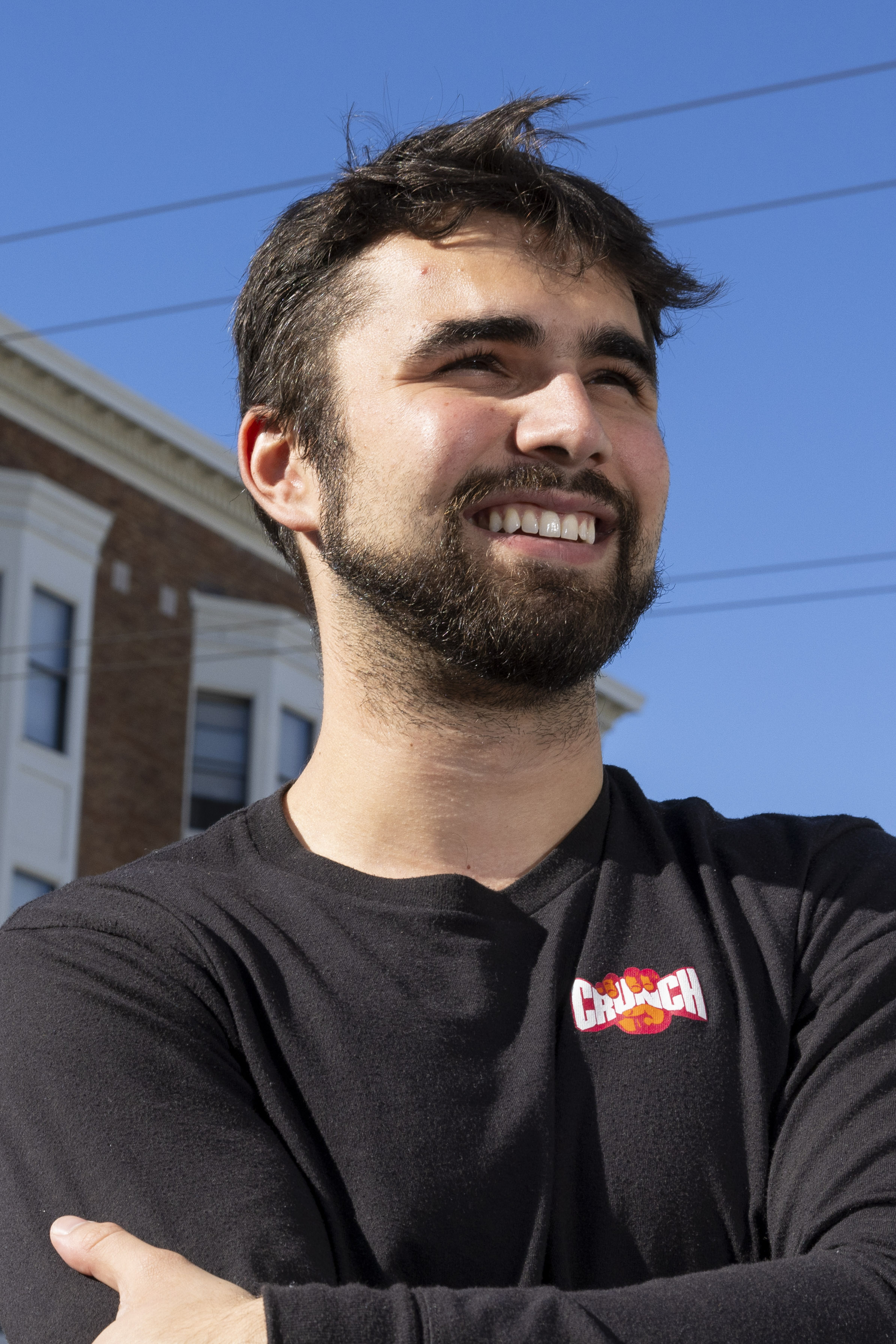 A man with a beard smiles while standing outdoors. He is wearing a black shirt with a colorful &quot;Crunch&quot; logo. In the background, there's a brick building.