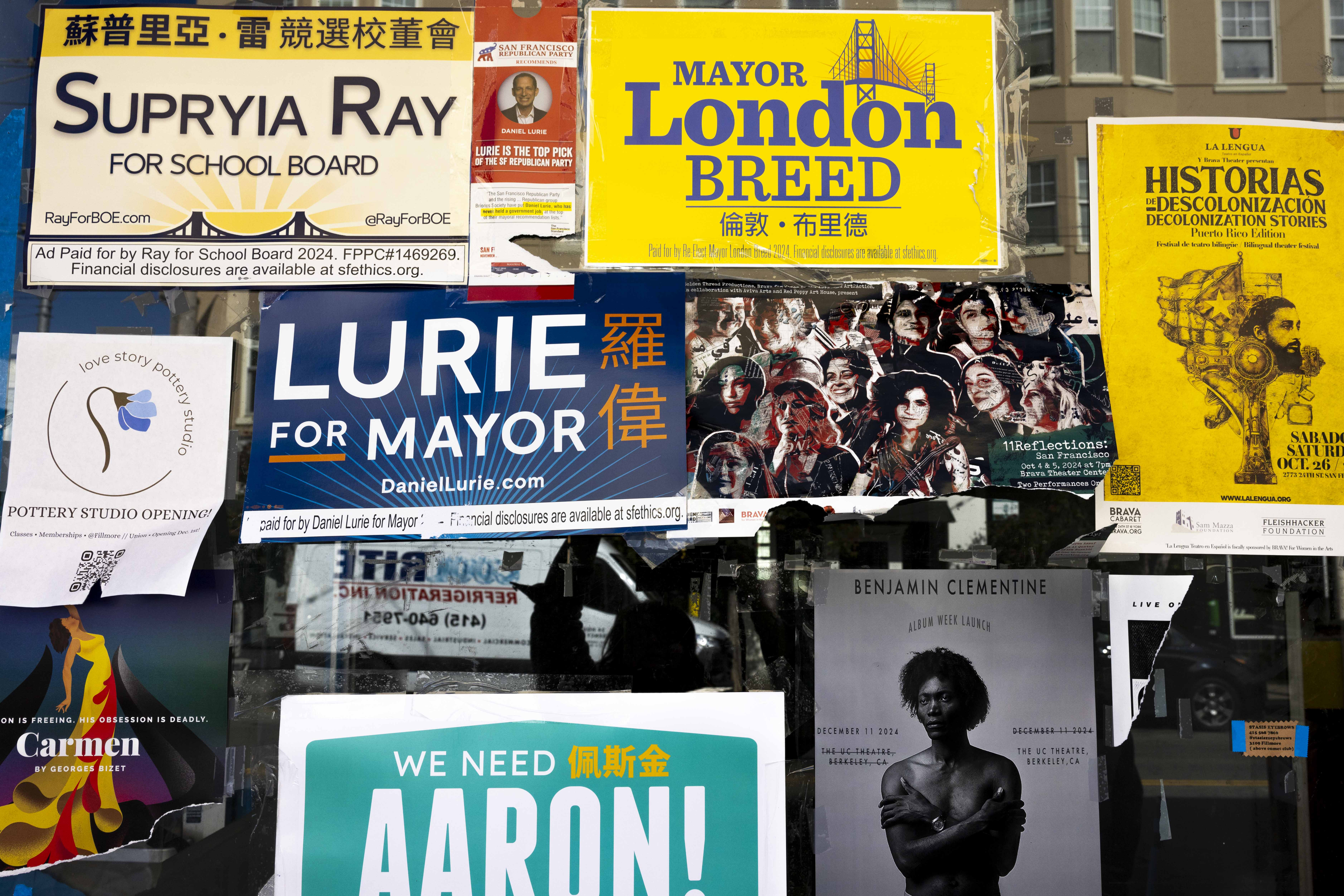 The image shows a collage of posters on a window, featuring political campaign ads, event flyers, and a Benjamin Clementine concert poster.