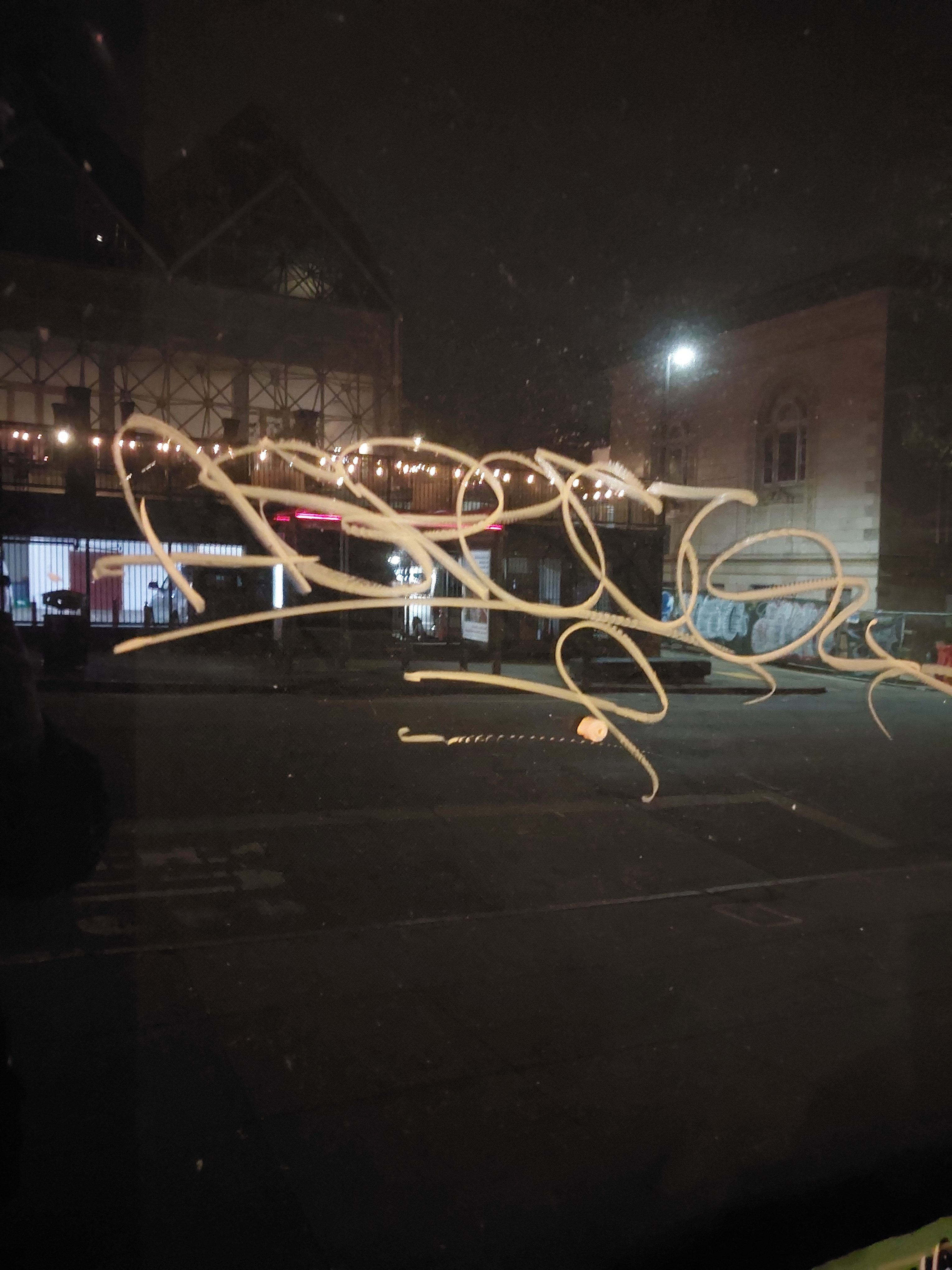 The image shows a night scene with a dimly lit street and a building. The window has a squiggly pattern drawn on it, and there are string lights hanging outside.