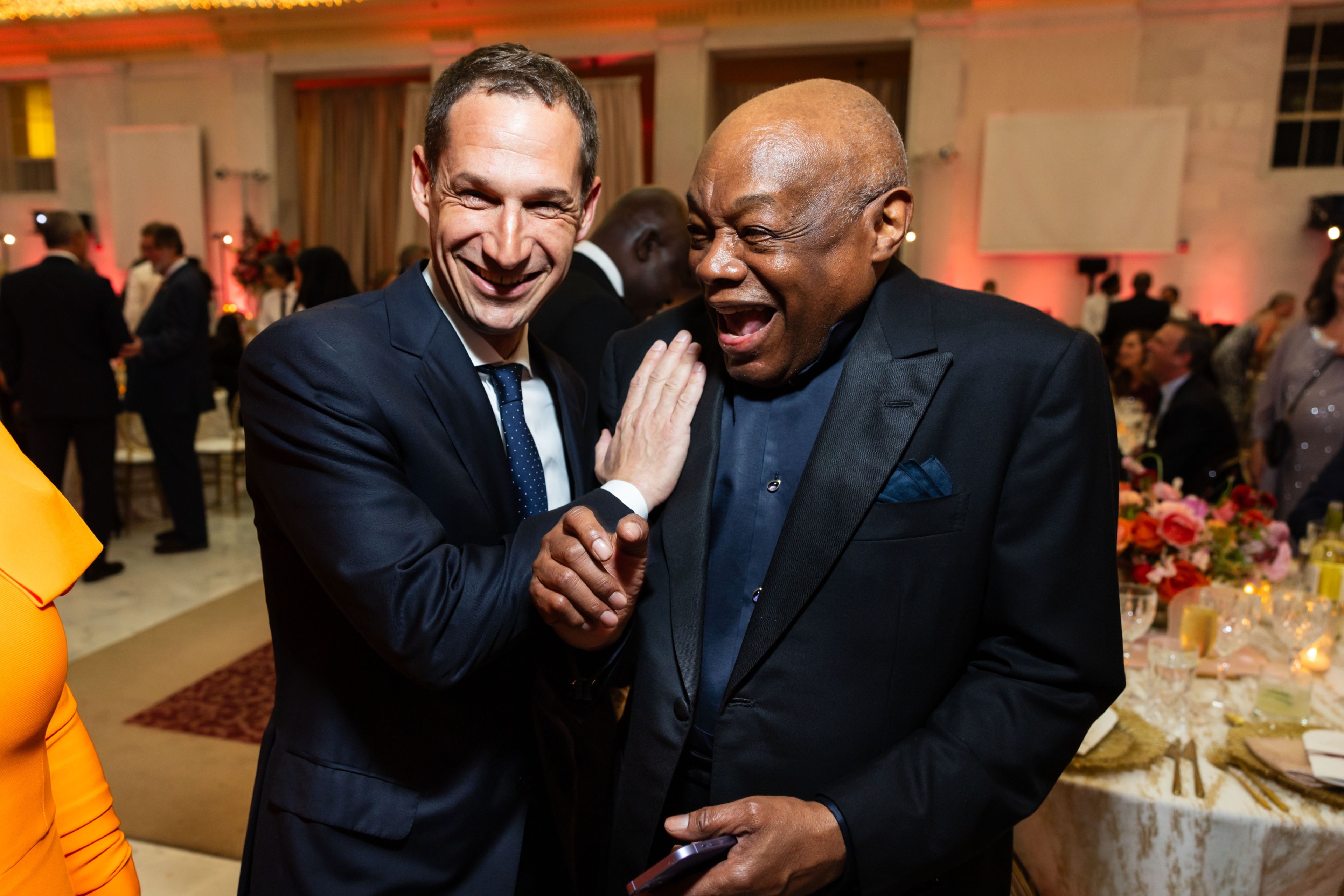 Two men in suits are laughing and embracing at a lively indoor event, surrounded by people and tables adorned with colorful flowers and candles.