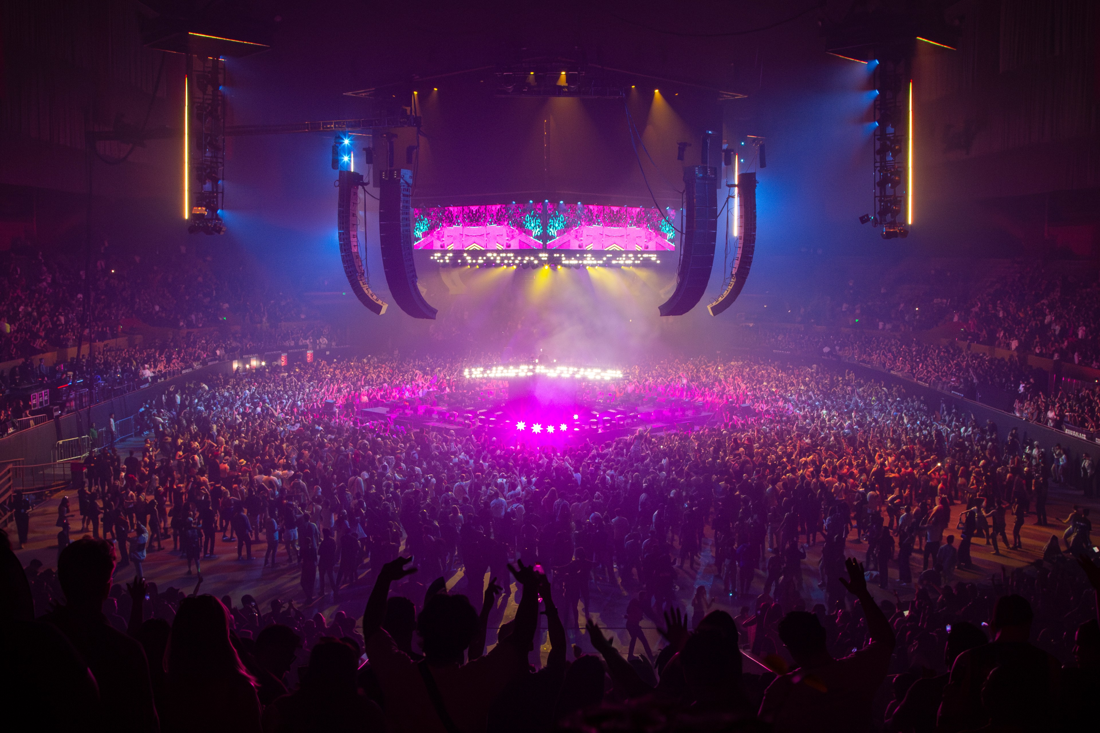 A crowded concert venue features a brightly lit, circular stage surrounded by an audience. Colorful lights and large speakers create an energetic atmosphere.