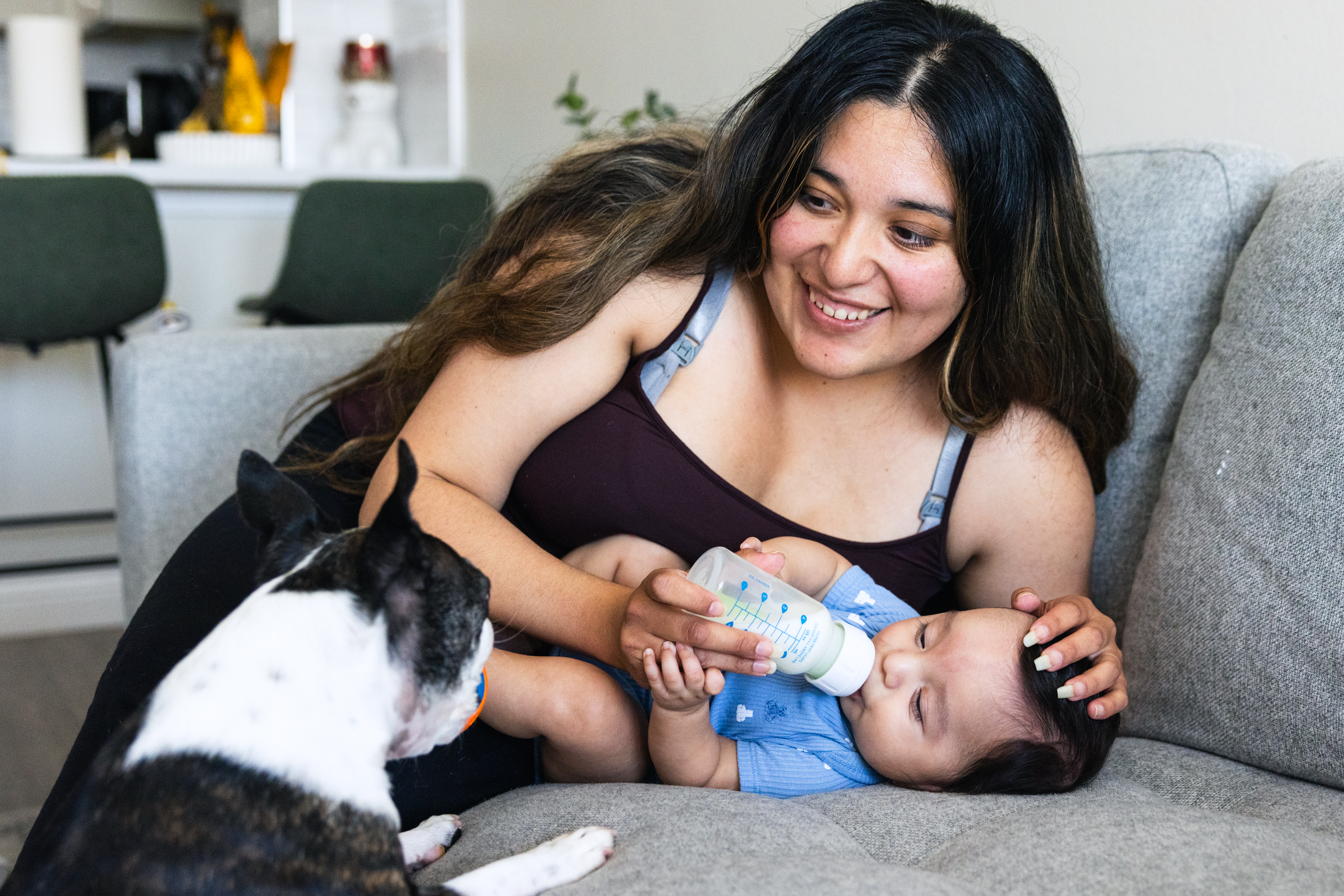 A woman smiles as she feeds a baby with a bottle while lying on a gray couch. A small black-and-white dog sits nearby, looking at them.