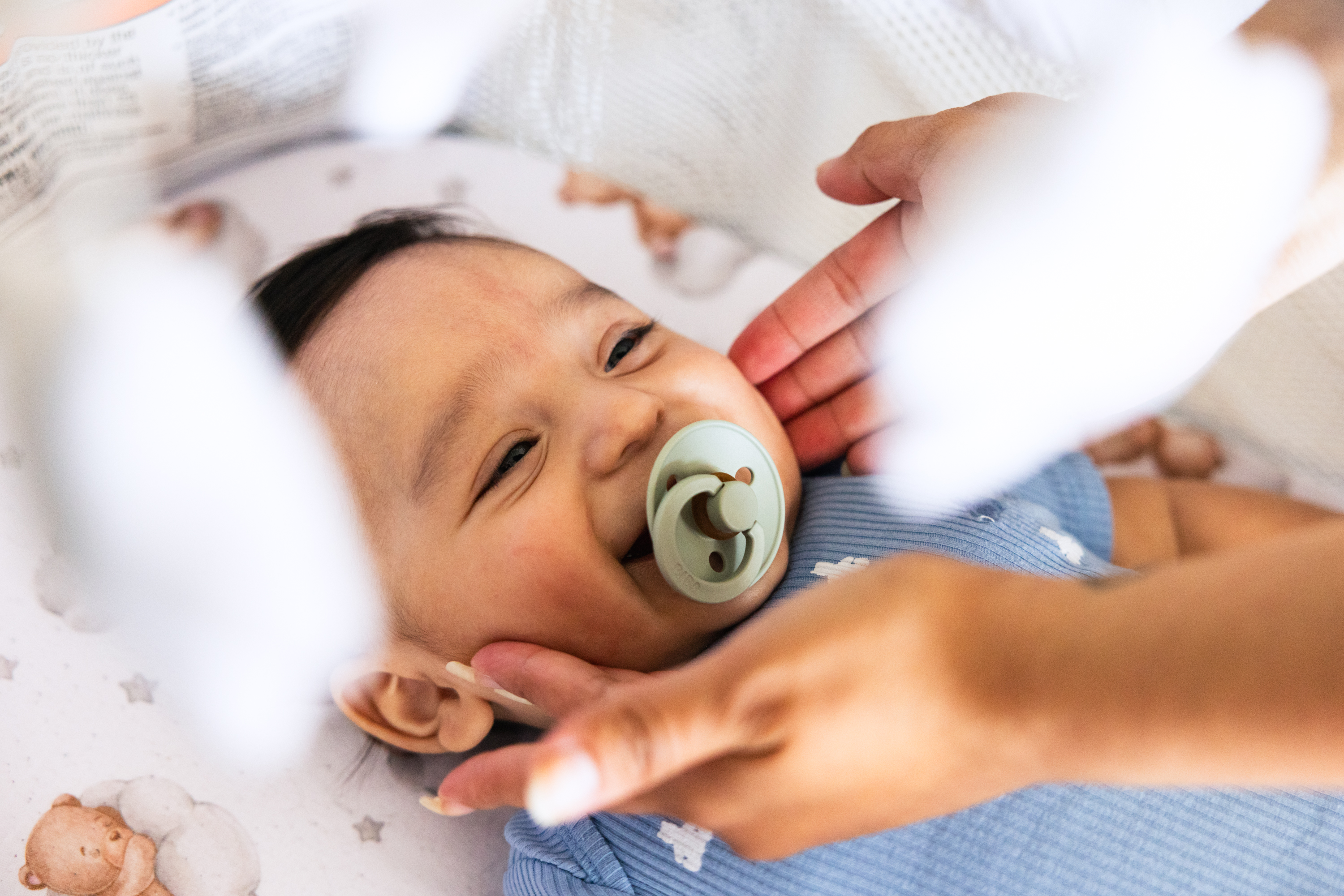 A baby with a pacifier smiles while lying on a patterned blanket. An adult's hands gently cradle the baby's face, creating a warm, tender scene.