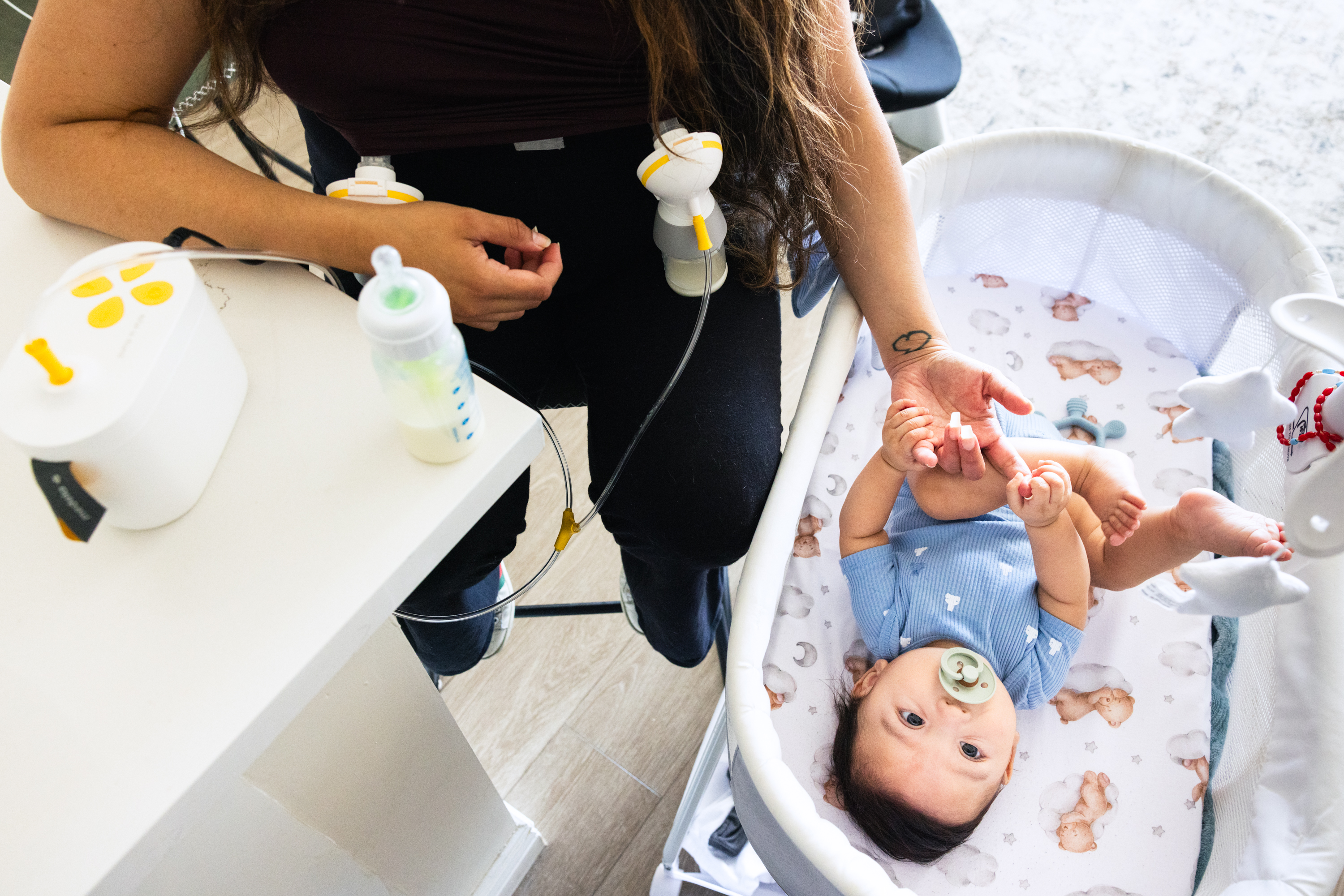 A woman is using a breast pump while seated, with a baby in a bassinet nearby. The baby has a pacifier and holds the woman's finger. A bottle is on the table.