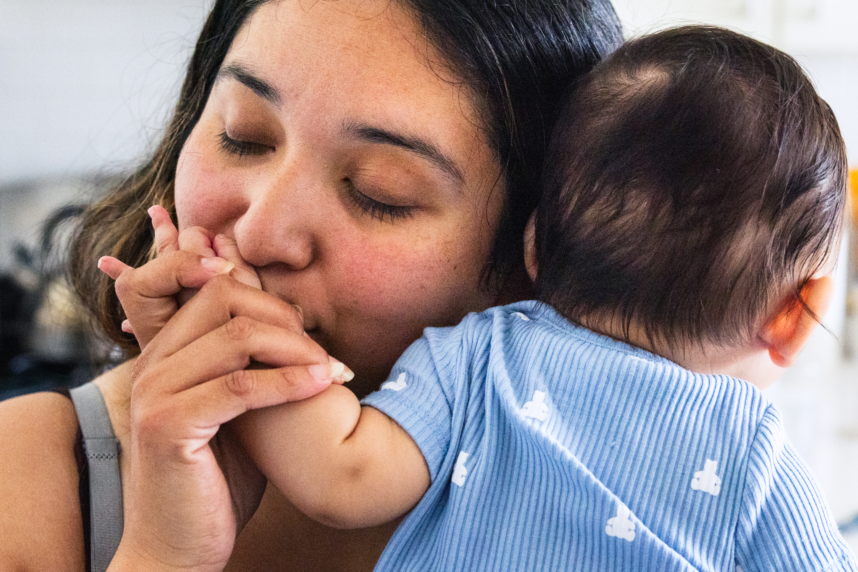 A woman with closed eyes kisses the hand of a baby she is holding against her shoulder. The baby is wearing a blue striped outfit with small white designs.