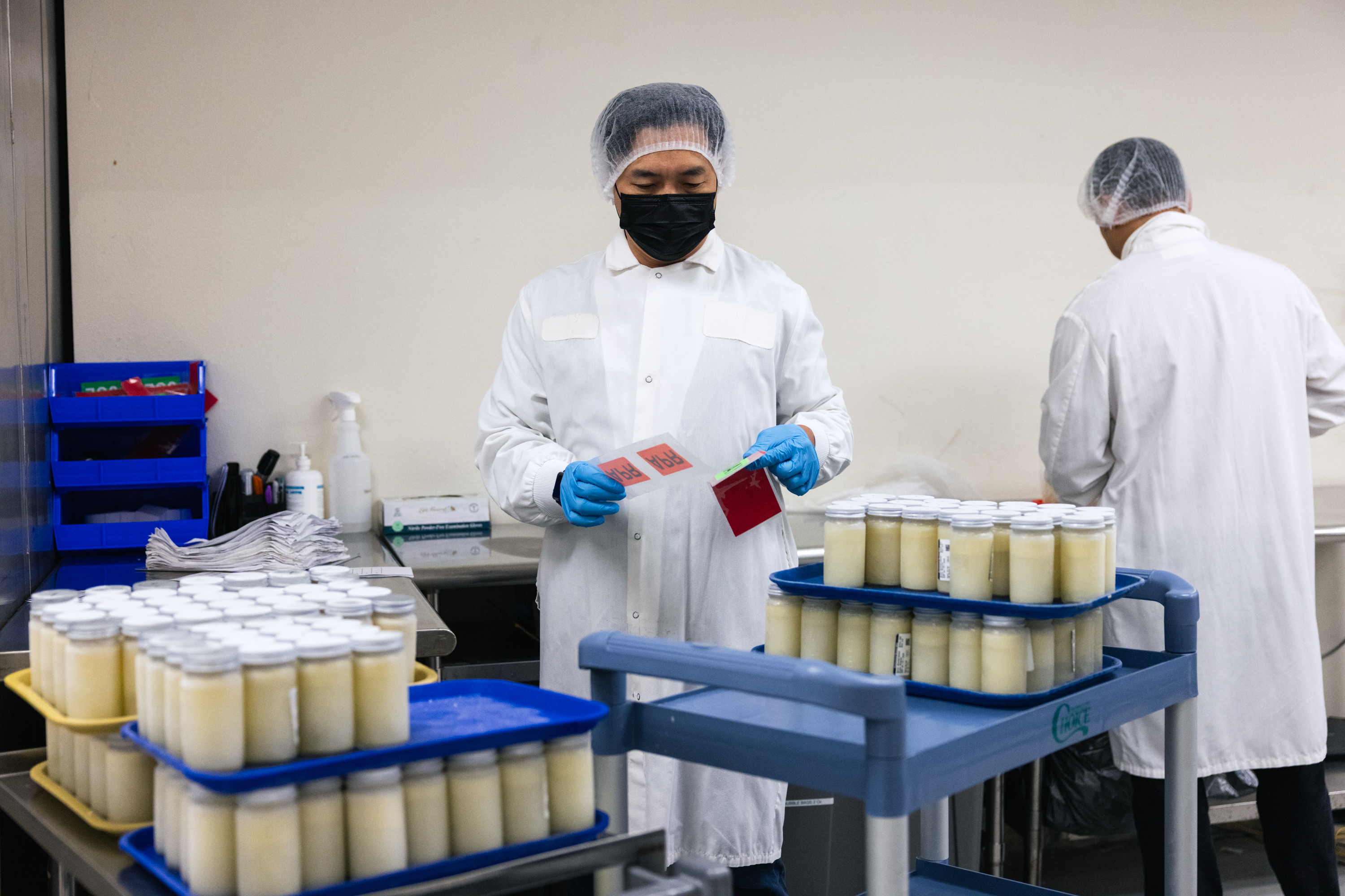 Two people in lab coats and hairnets work in a facility with trays of sealed containers on carts, wearing gloves and masks, handling labels.