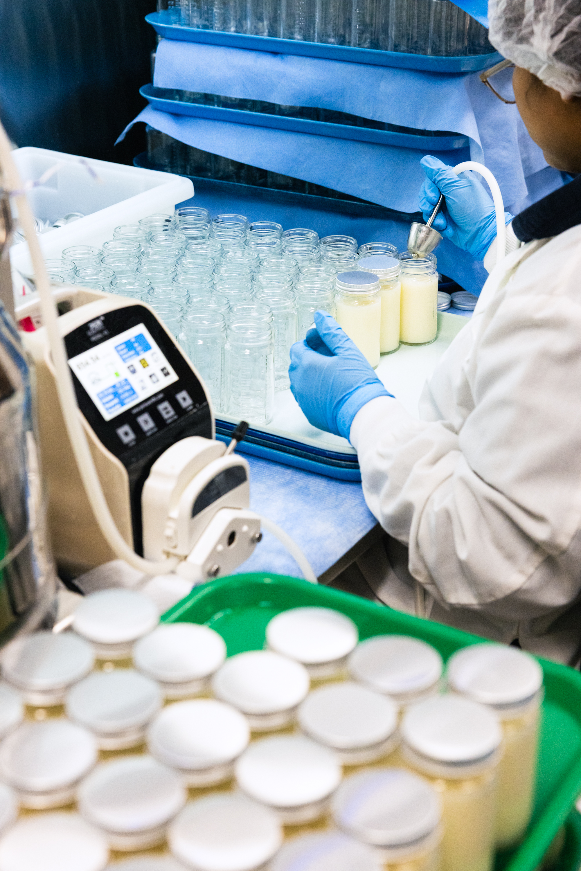 A person in a lab coat and gloves fills jars with a creamy substance from a machine. Empty jars and sealed lids are also visible on the counter.