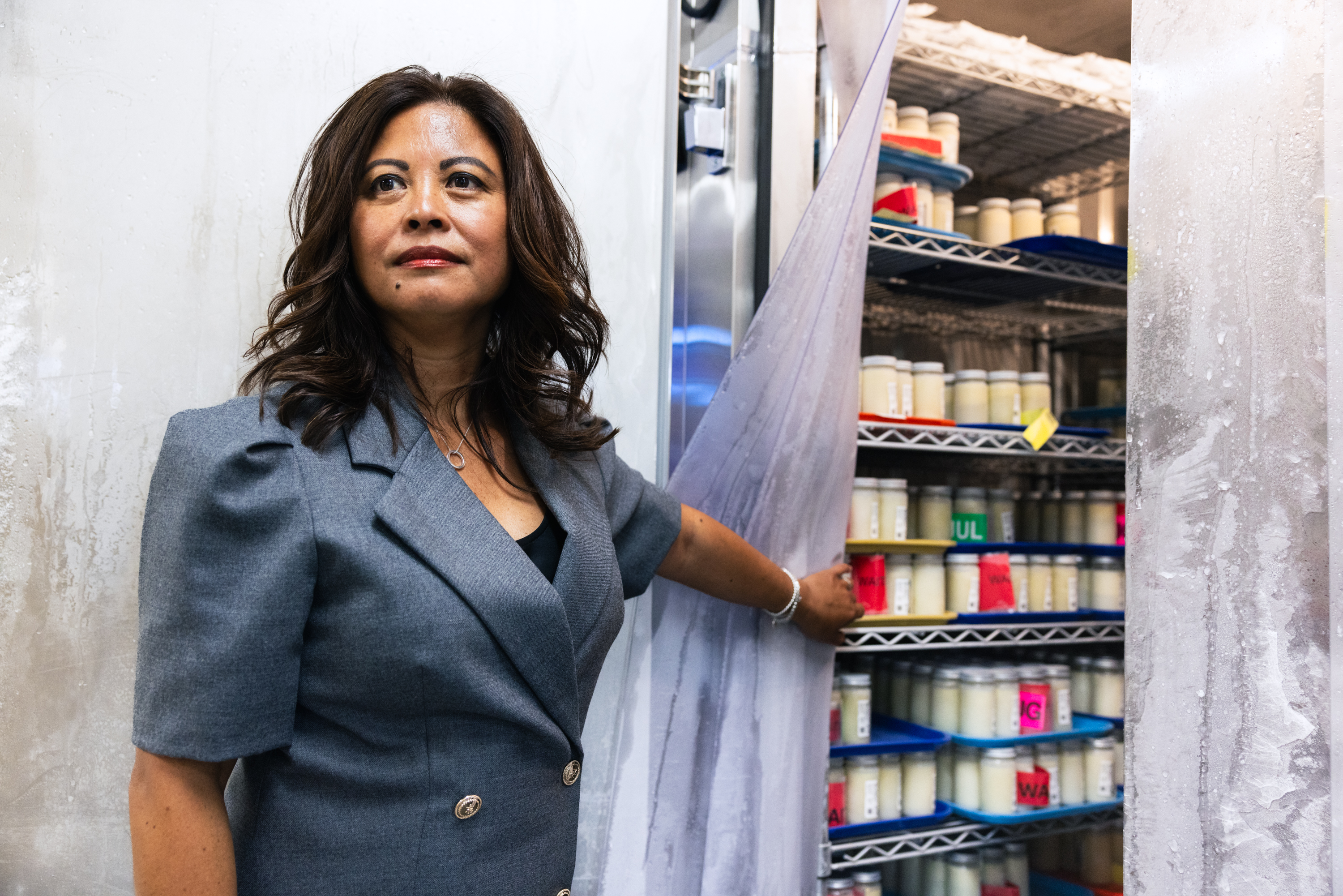 A woman in a gray blazer stands by an open storage area filled with shelves of jars. She holds a plastic curtain aside, revealing neatly arranged containers.