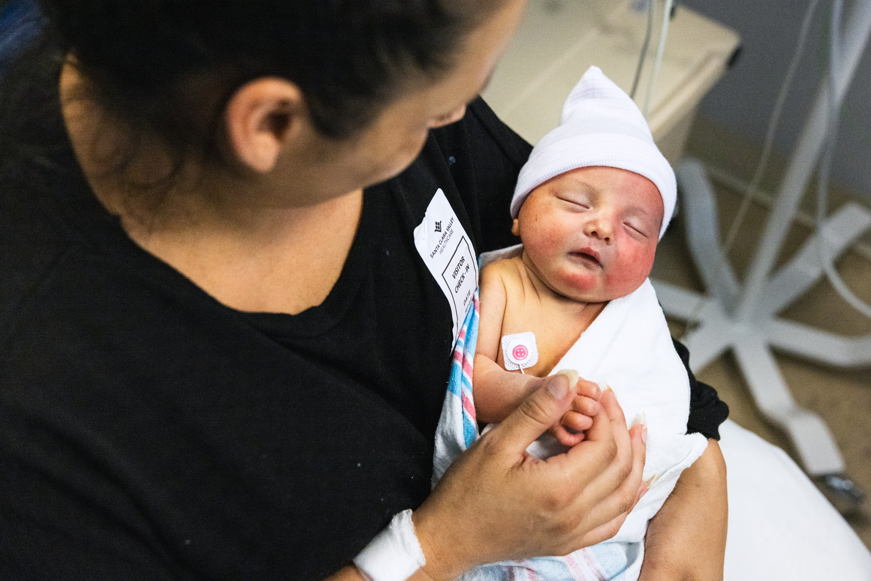 A woman gently cradles a sleeping newborn wrapped in a blanket, wearing a white cap. The woman is holding the baby's hand, conveying tenderness and care.