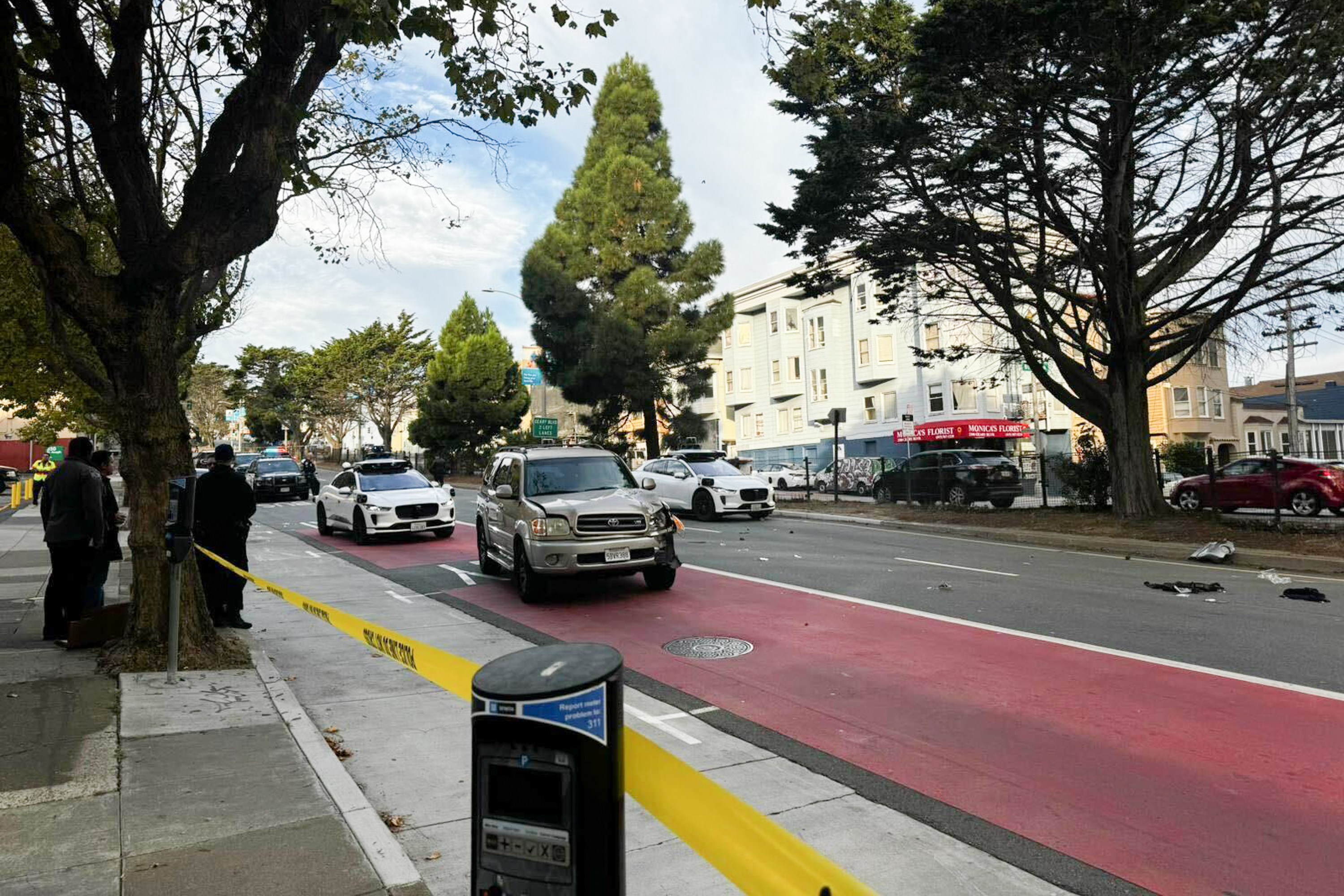 A street scene shows cars on a road with a red lane, surrounded by trees and buildings. Police tape blocks off an area, with people standing on the sidewalk. Debris is scattered.