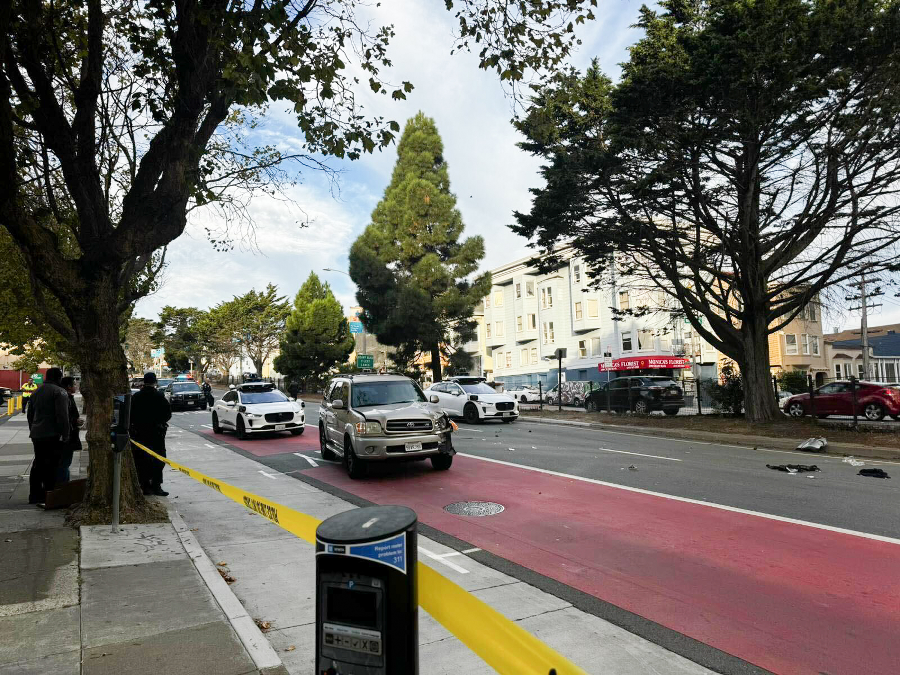 A street scene shows cars on a road with a red lane, surrounded by trees and buildings. Police tape blocks off an area, with people standing on the sidewalk. Debris is scattered.