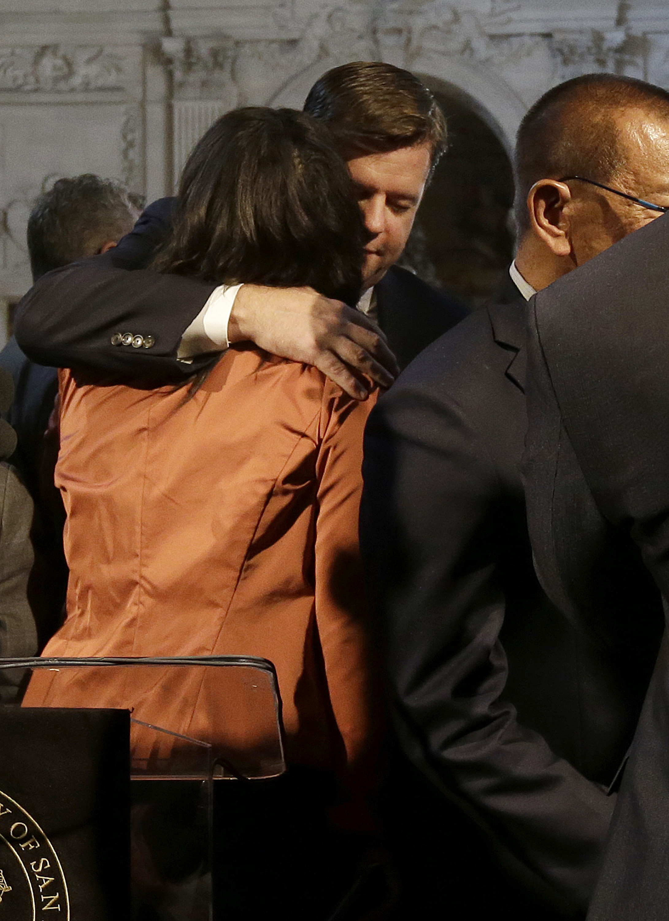 Mark Farrell faces the camera, and hugs London Breed, whose back is turned to the frame. they are standing in city hall