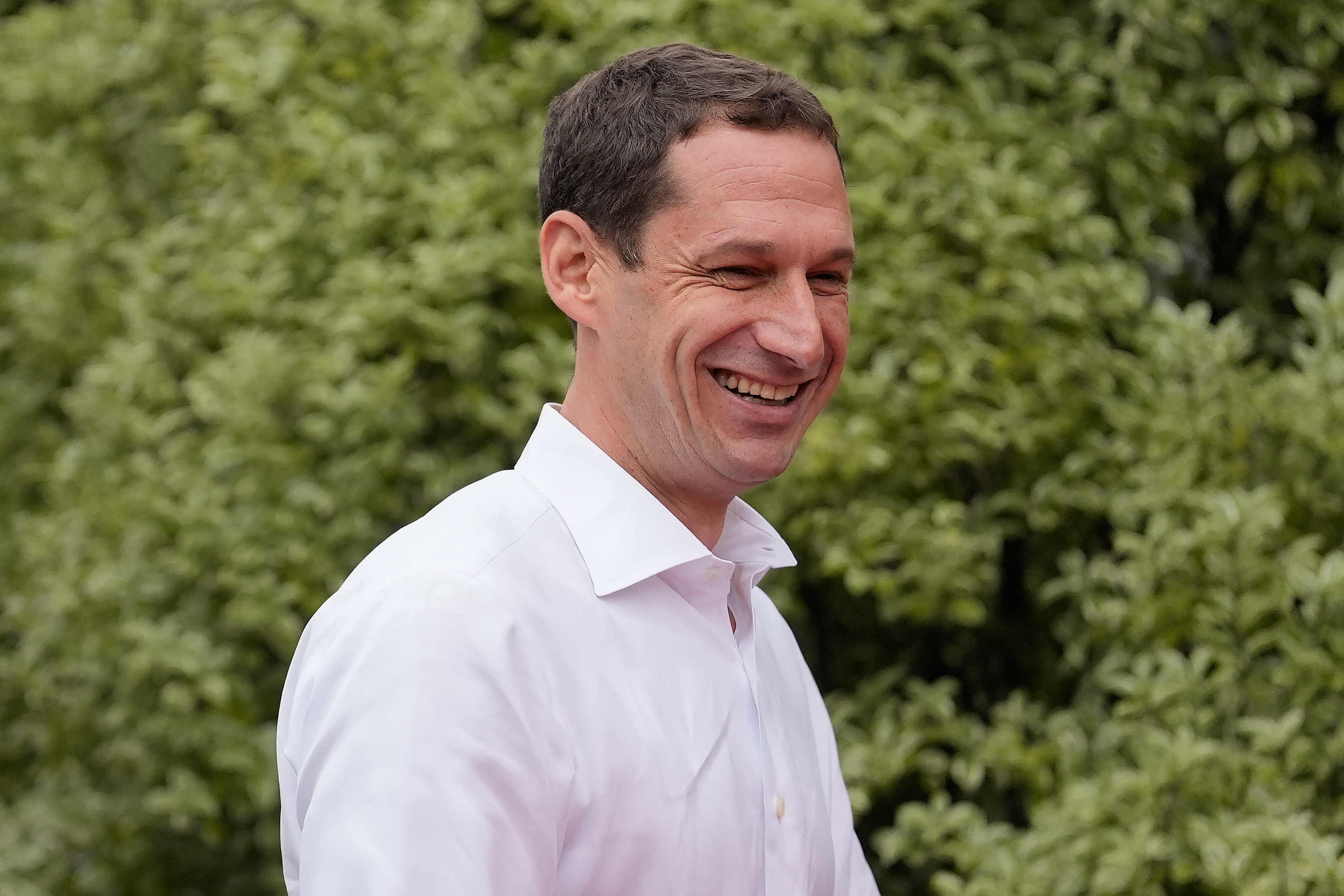 A man in a white shirt is smiling in front of lush green foliage.