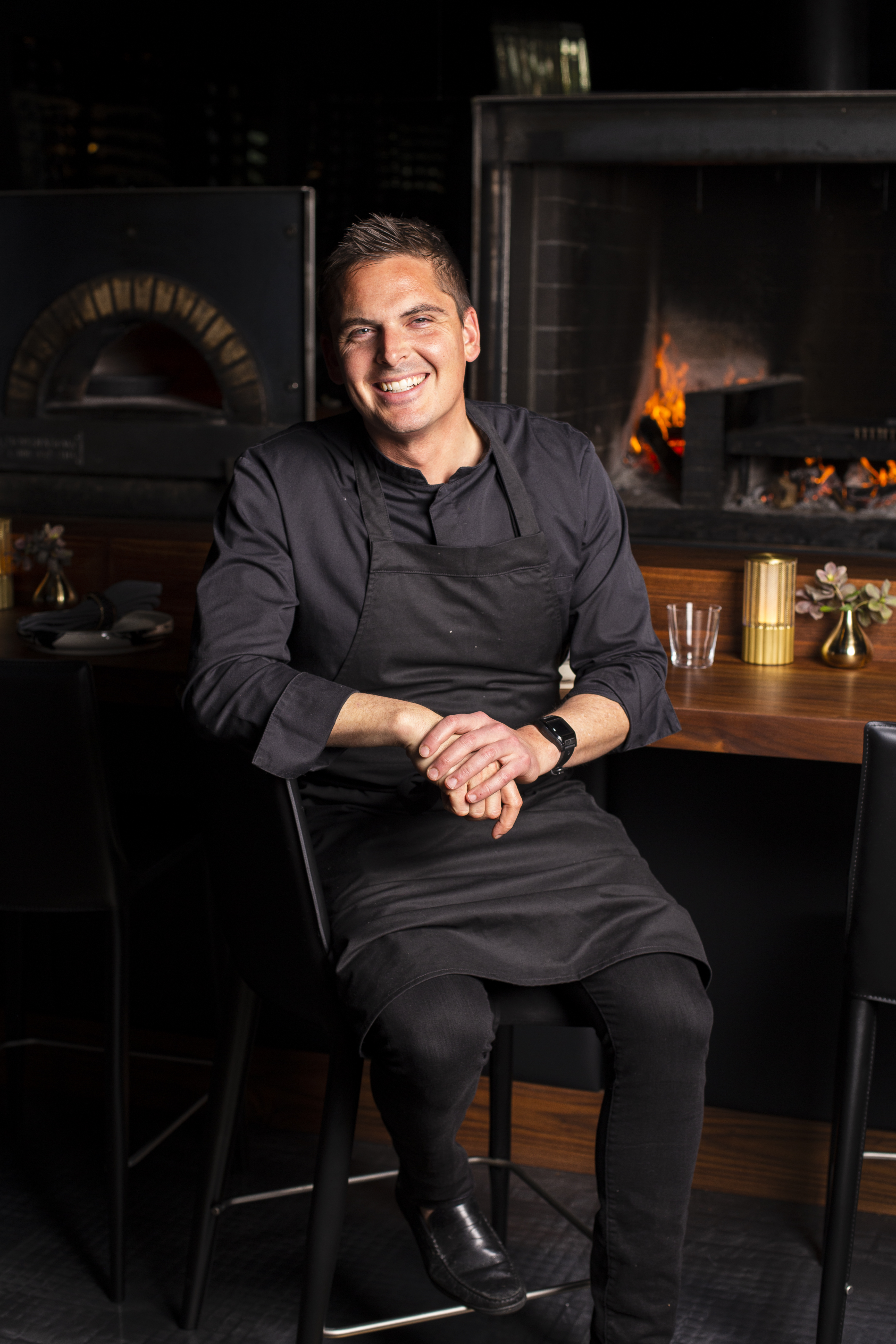 A smiling man in black chef attire sits in front of a wood-fired oven, with a fire burning. He rests his hands on his lap, creating a welcoming atmosphere.