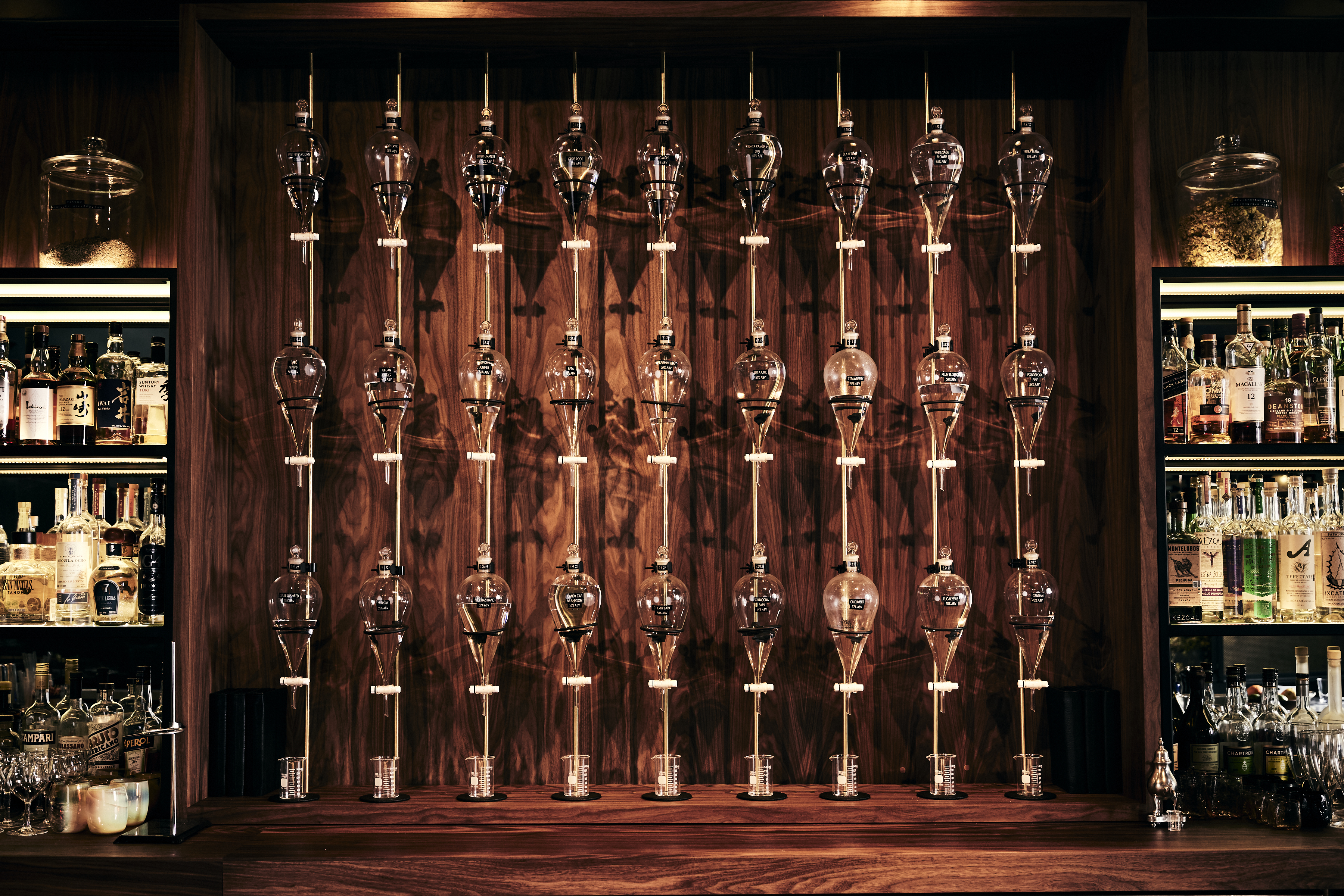 A wall-mounted coffee drip setup with multiple glass vessels lined up. Shelves on either side hold various liquor bottles and glasses against a wooden backdrop.