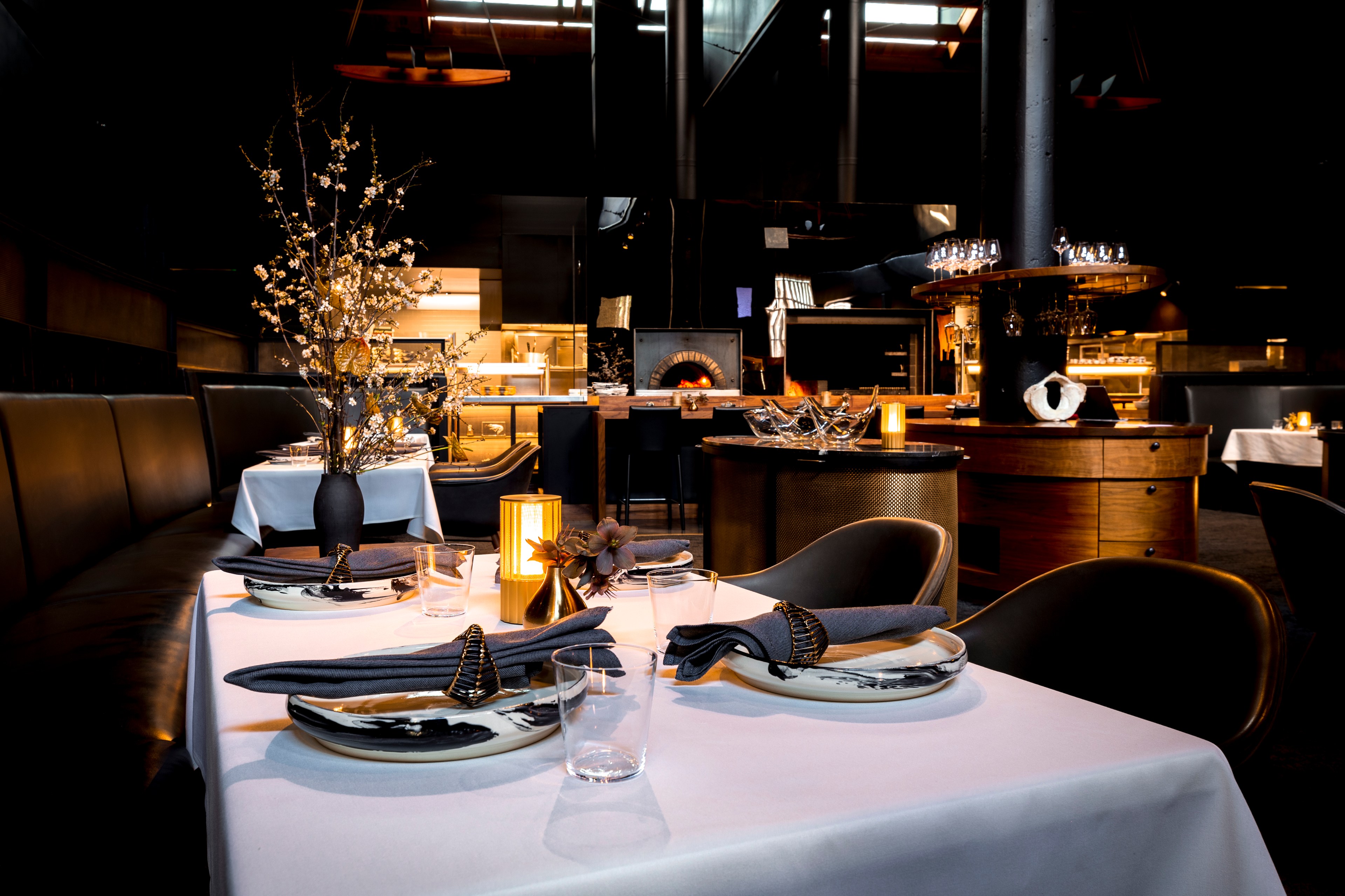 A stylish restaurant interior features elegantly set tables with white cloths, decorative plates, folded napkins, and branches in vases, lit by warm ambient lighting.