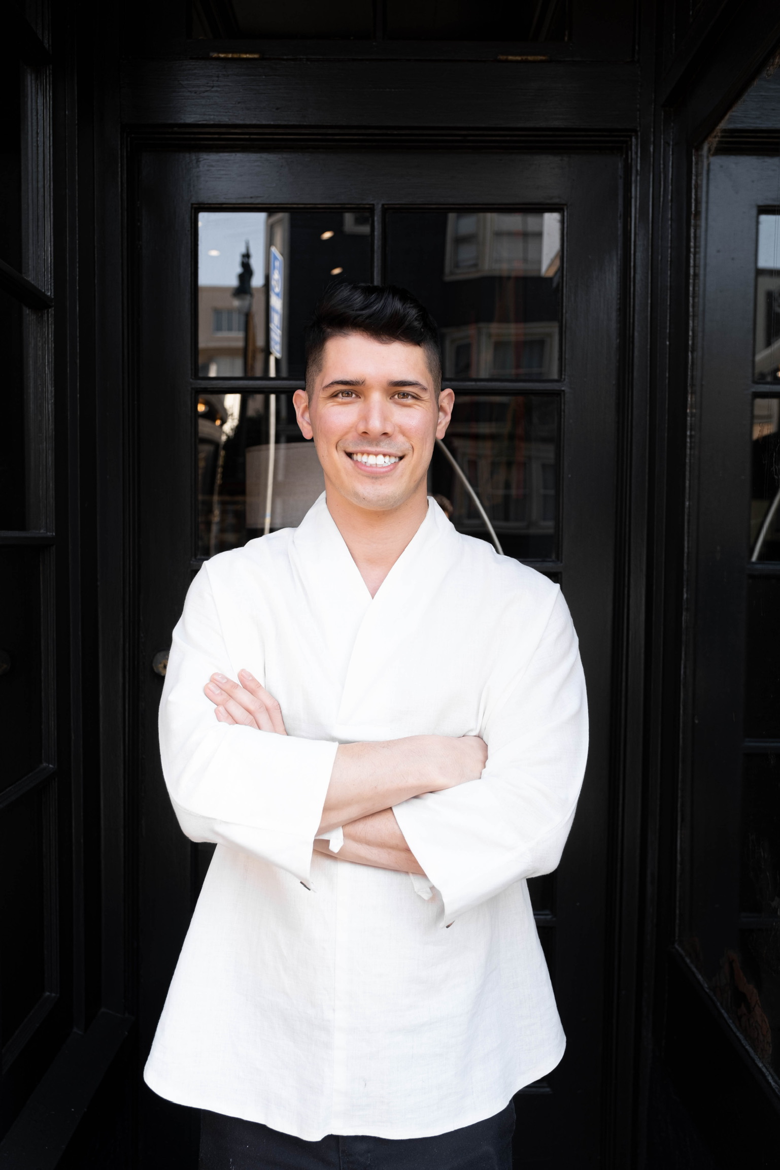 A person stands smiling with arms crossed, wearing a white chef's coat, in front of a dark wooden door with glass panes.