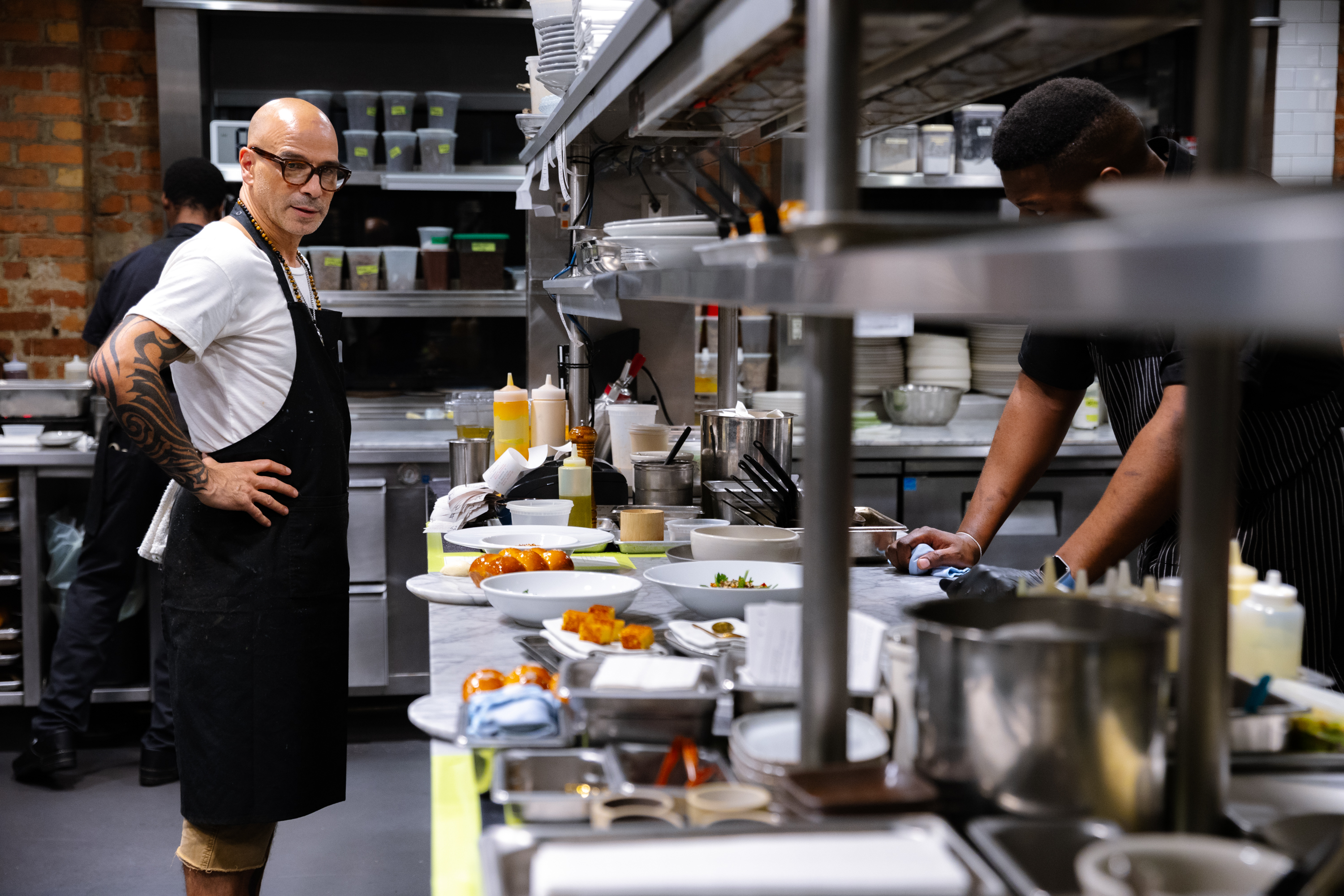 Two chefs work in a bustling kitchen; one stands with hands on hips, the other prepares food at a counter. The space is filled with utensils and ingredients.