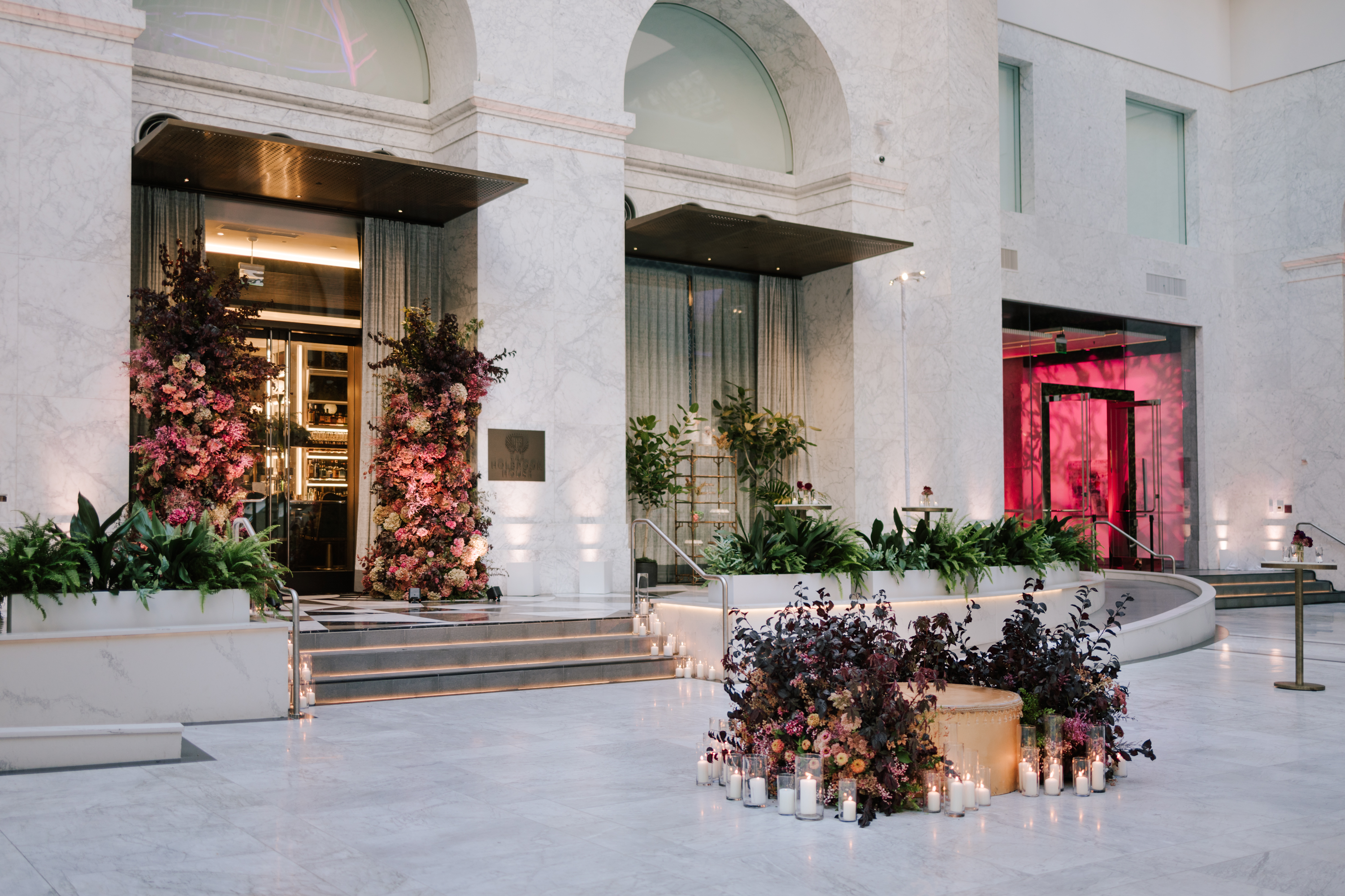 A grand marble entrance features lush floral arrangements and numerous candles, leading up stairs to a building with large arched windows and curtains.