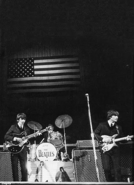 The image shows a black-and-white photo of The Beatles performing on stage with instruments. An American flag hangs above, and &quot;The Beatles&quot; is on the drum kit.