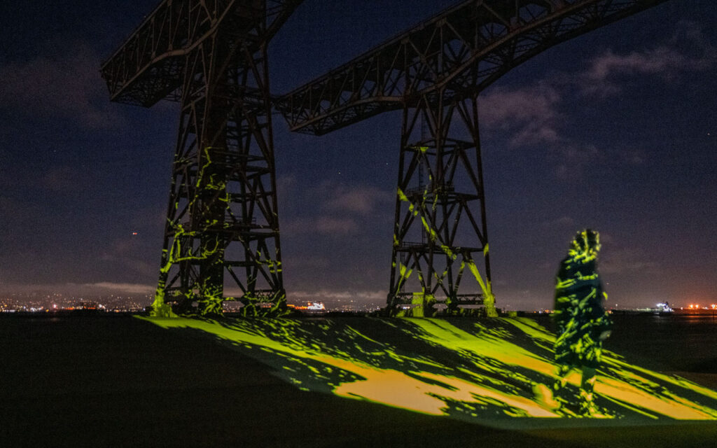 Two large metal structures stand against a night sky. Yellow-green lights project patterns on them and the ground, with distant city lights visible.