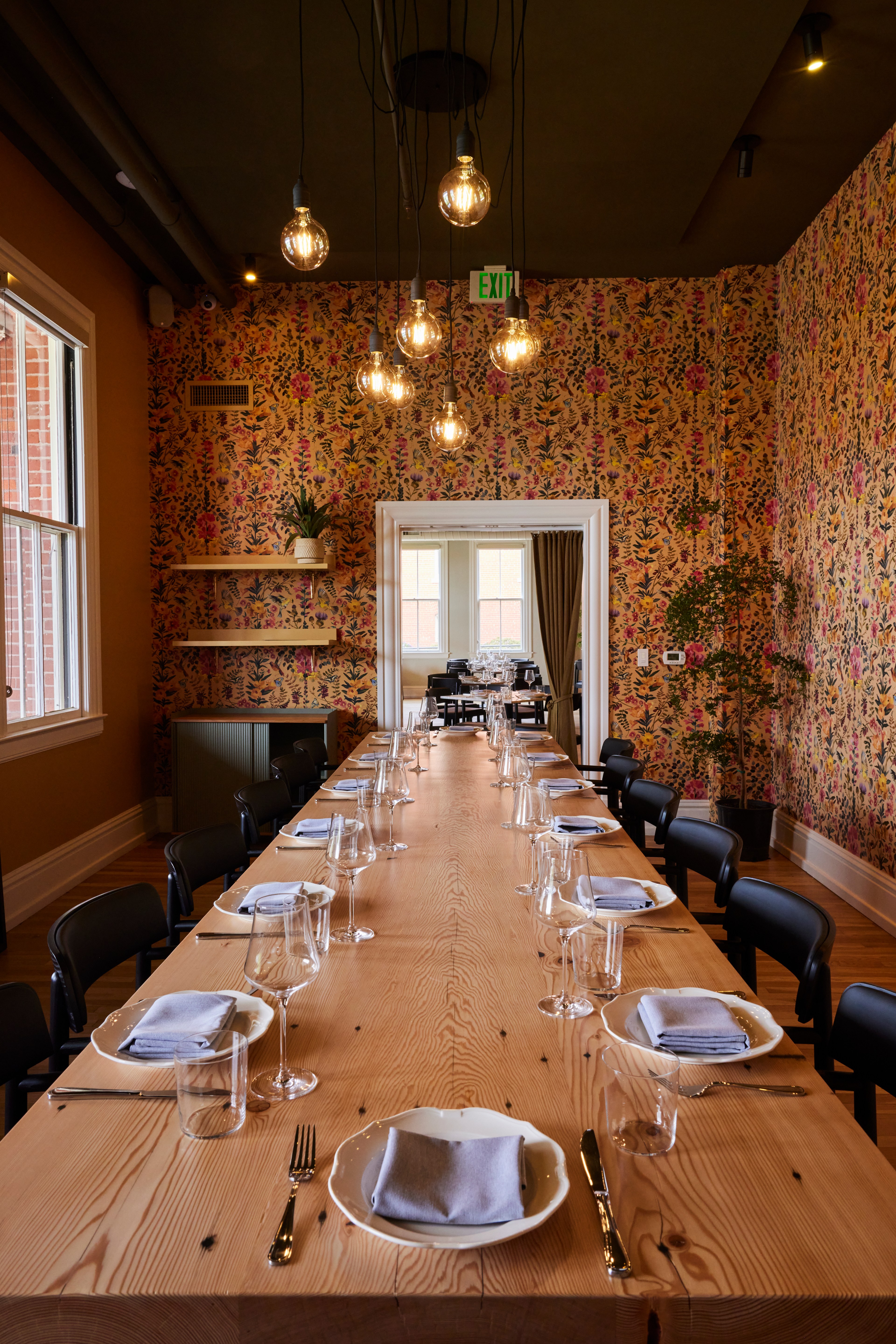 A long wooden table is set with plates, glasses, and napkins in a room with floral wallpaper and hanging bulbs. There is a window and a plant on the side.