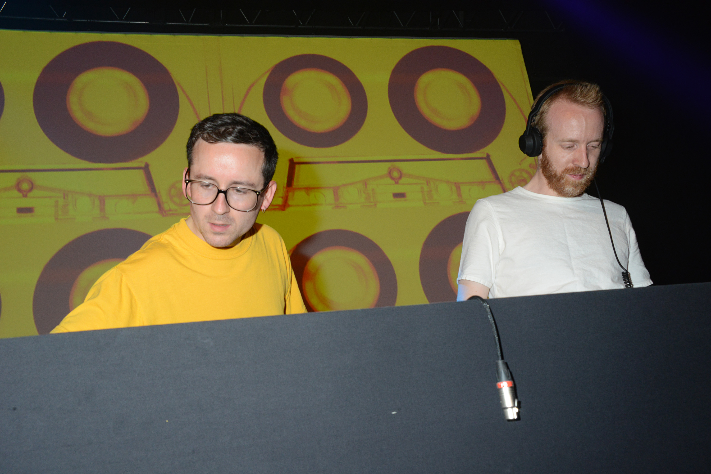 Two DJs are performing on stage. One wears a yellow shirt, the other a white shirt with headphones. A graphic of circles is displayed behind them.