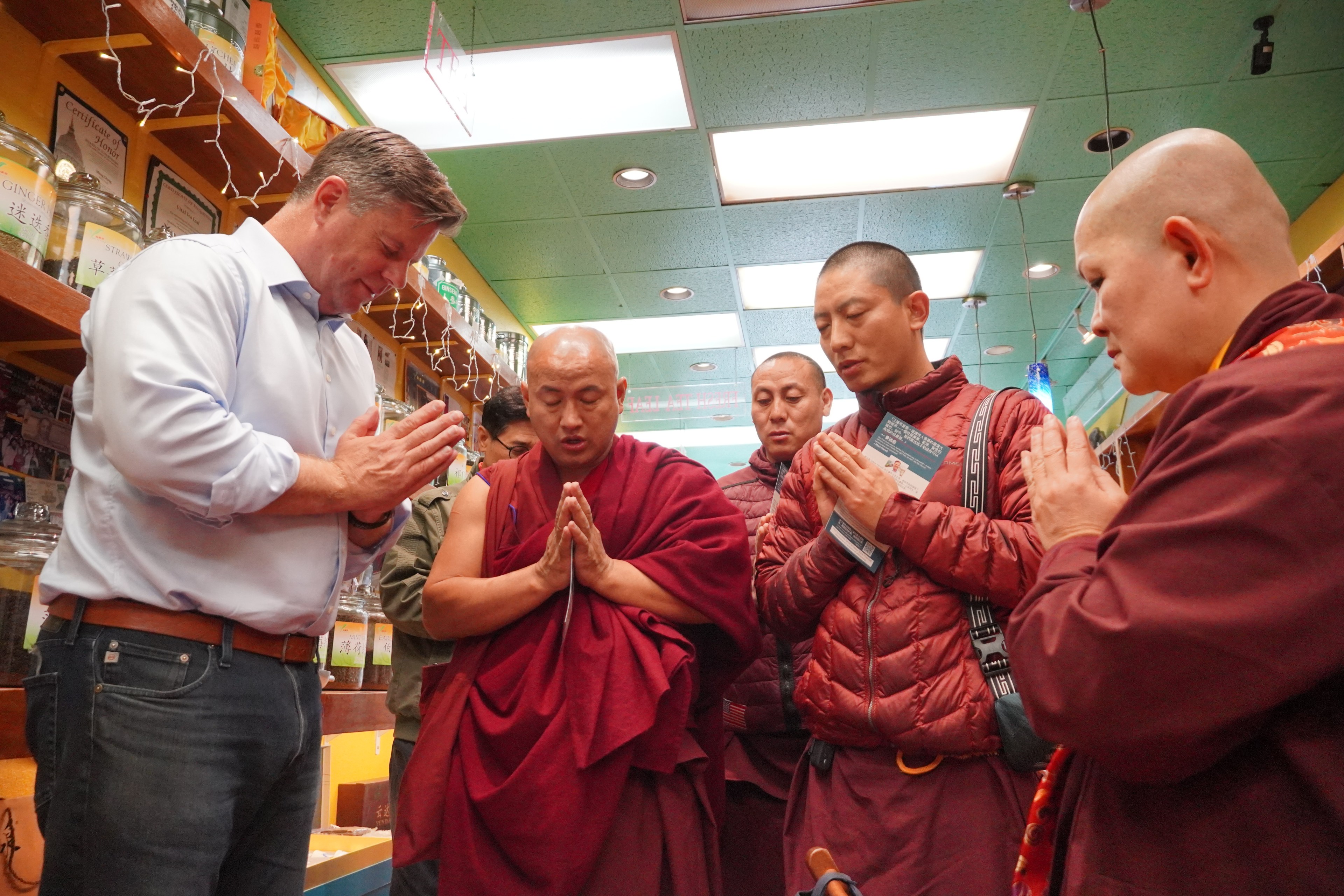 A group of five people, including Buddhist monks with shaved heads, stand in prayer with palms together inside a shop decorated with string lights.