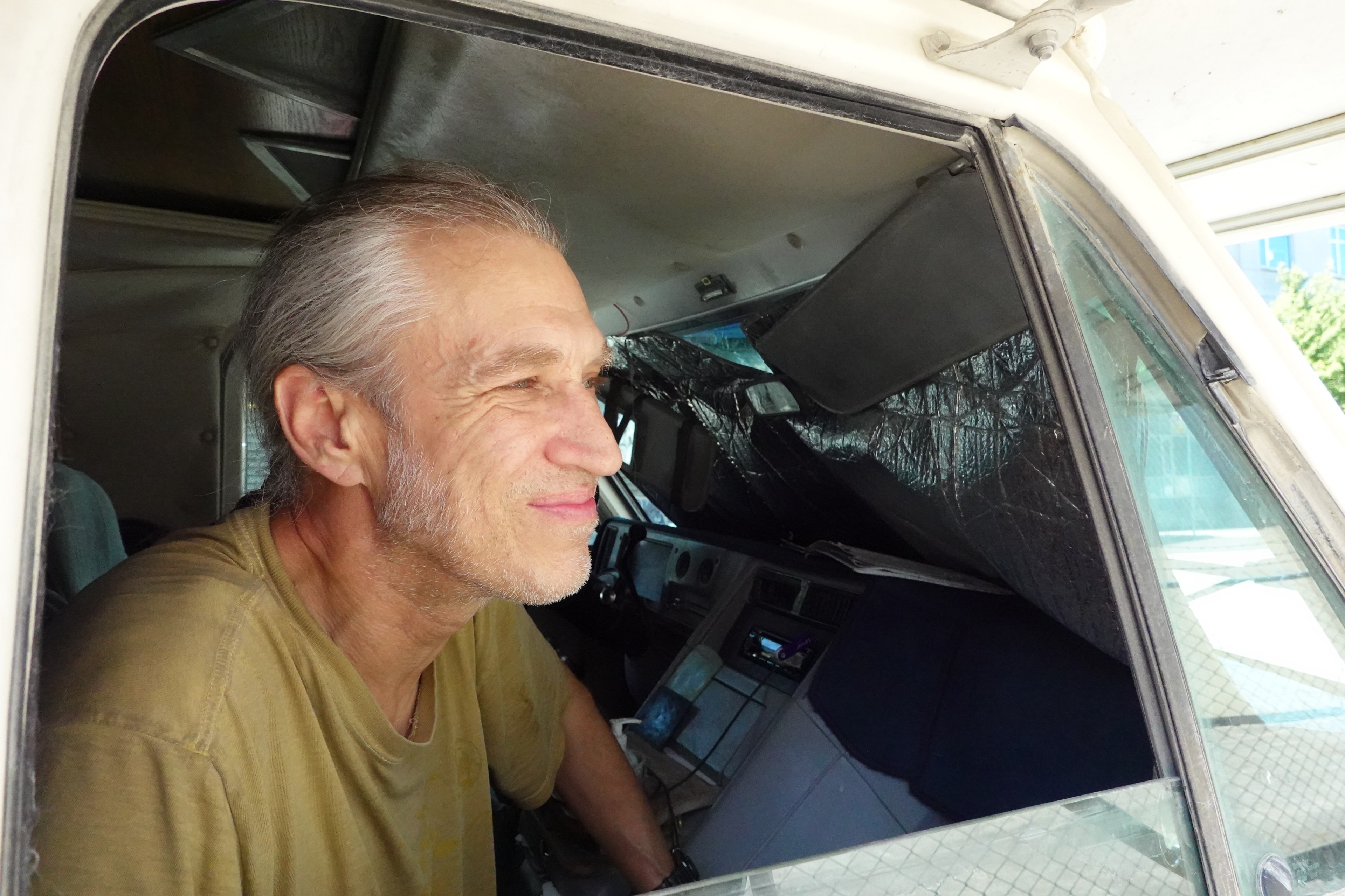 A man with gray hair is smiling while leaning out of a vehicle window. He wears a tan shirt, and the vehicle's interior appears cluttered with items.