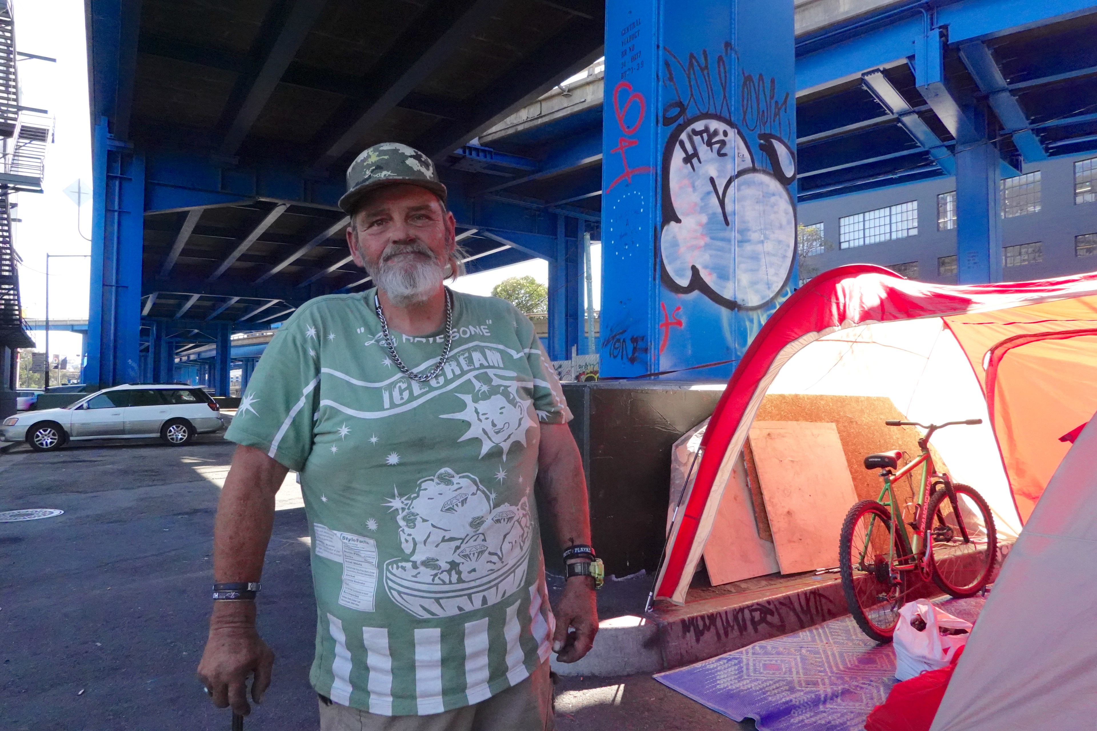 A man stands under a blue bridge next to a bright red-and-white tent. He's wearing a green ice cream-themed shirt and camo hat, with a bicycle nearby.