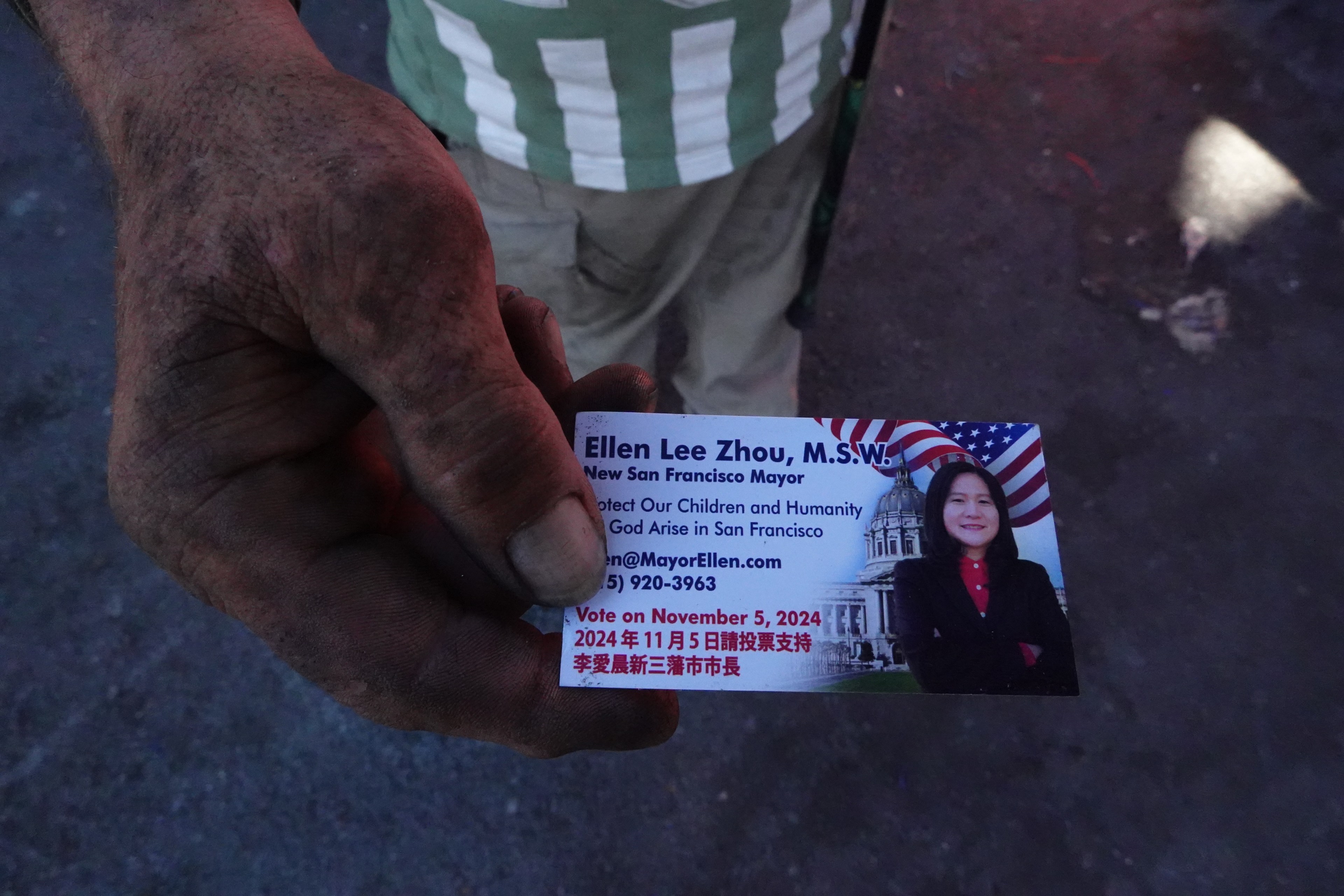 A dirty hand holds a campaign card for Ellen Lee Zhou, a San Francisco mayoral candidate, featuring her photo, contact info, and voting date on November 5, 2024.