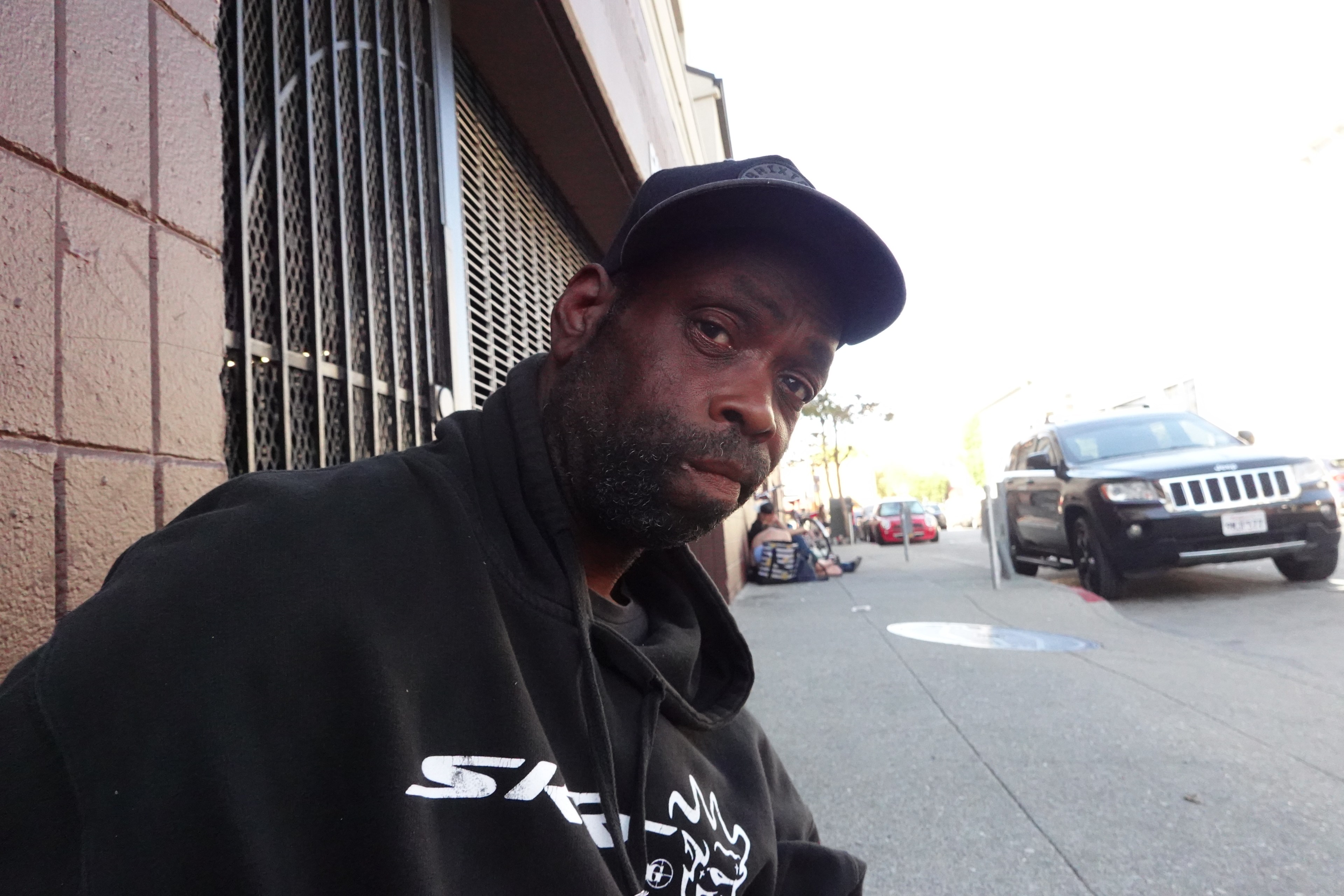 A man in a black hoodie and cap sits on a sidewalk by a building with metal grates. Cars and people are visible in the background.