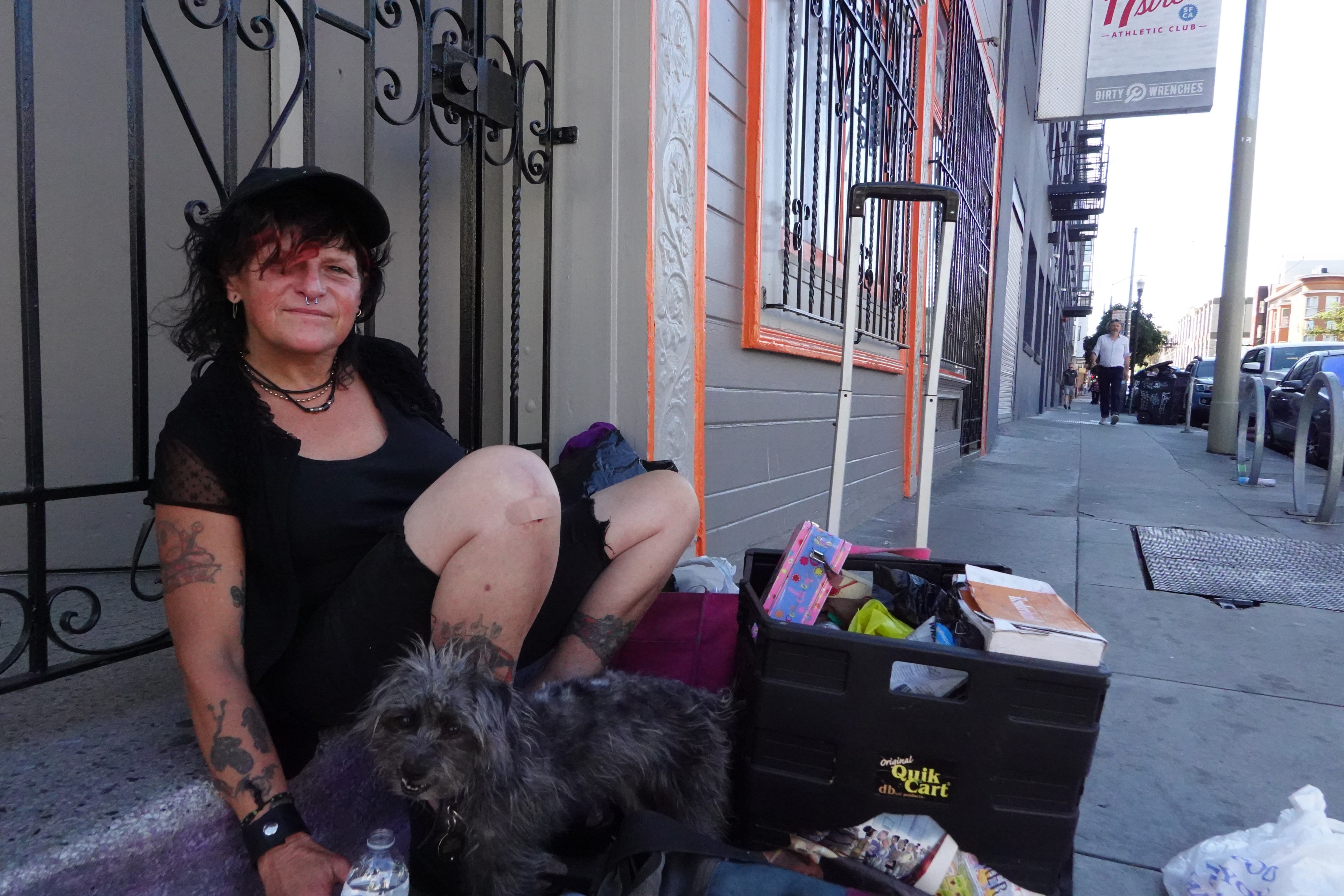 A person with tattoos and a nose ring sits on a sidewalk beside a small, shaggy dog. They have bags and a cart with various items nearby.