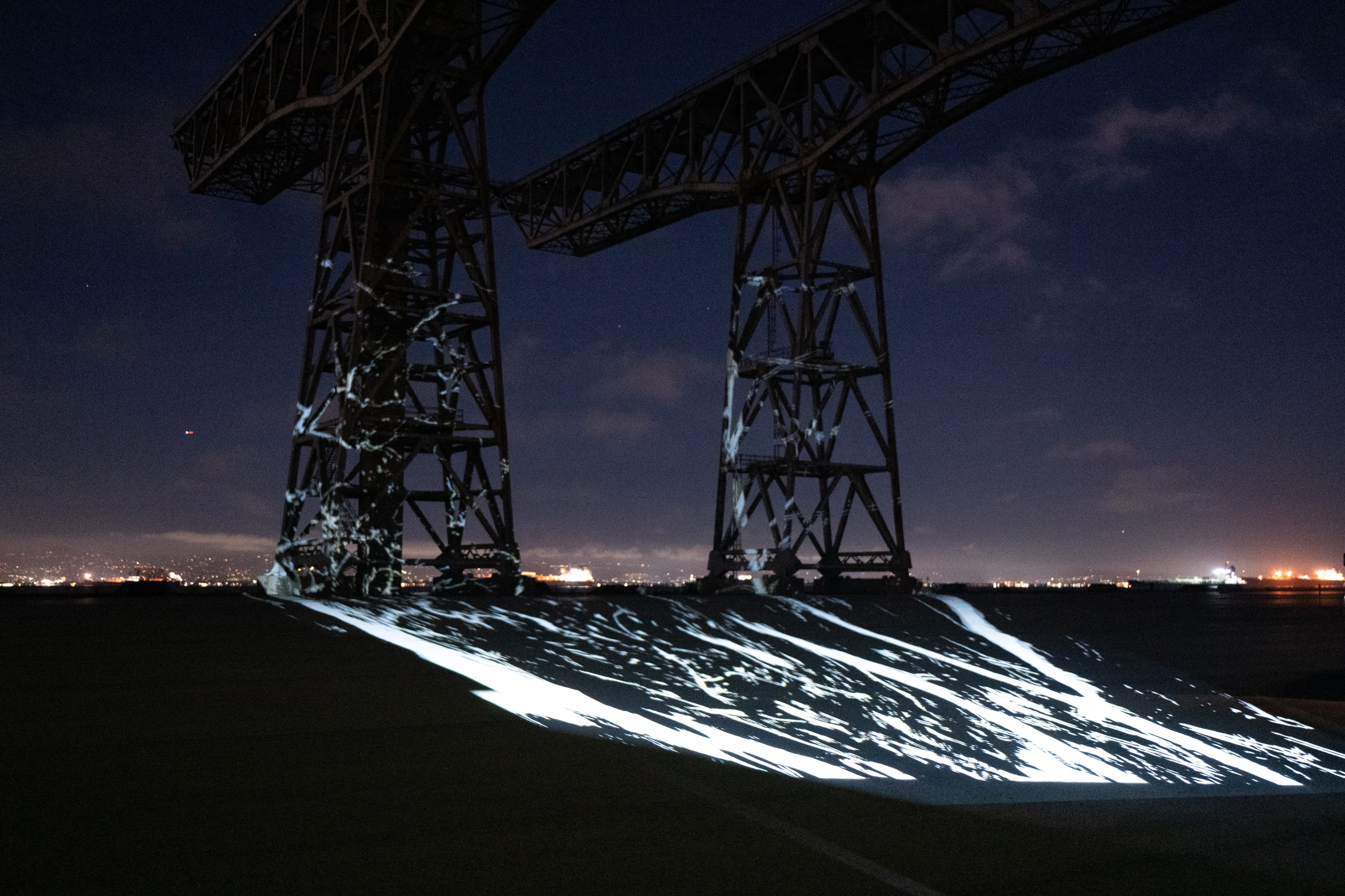 The image shows an illuminated industrial structure at night, with abstract light patterns projected on the ground and city lights twinkling in the distance.