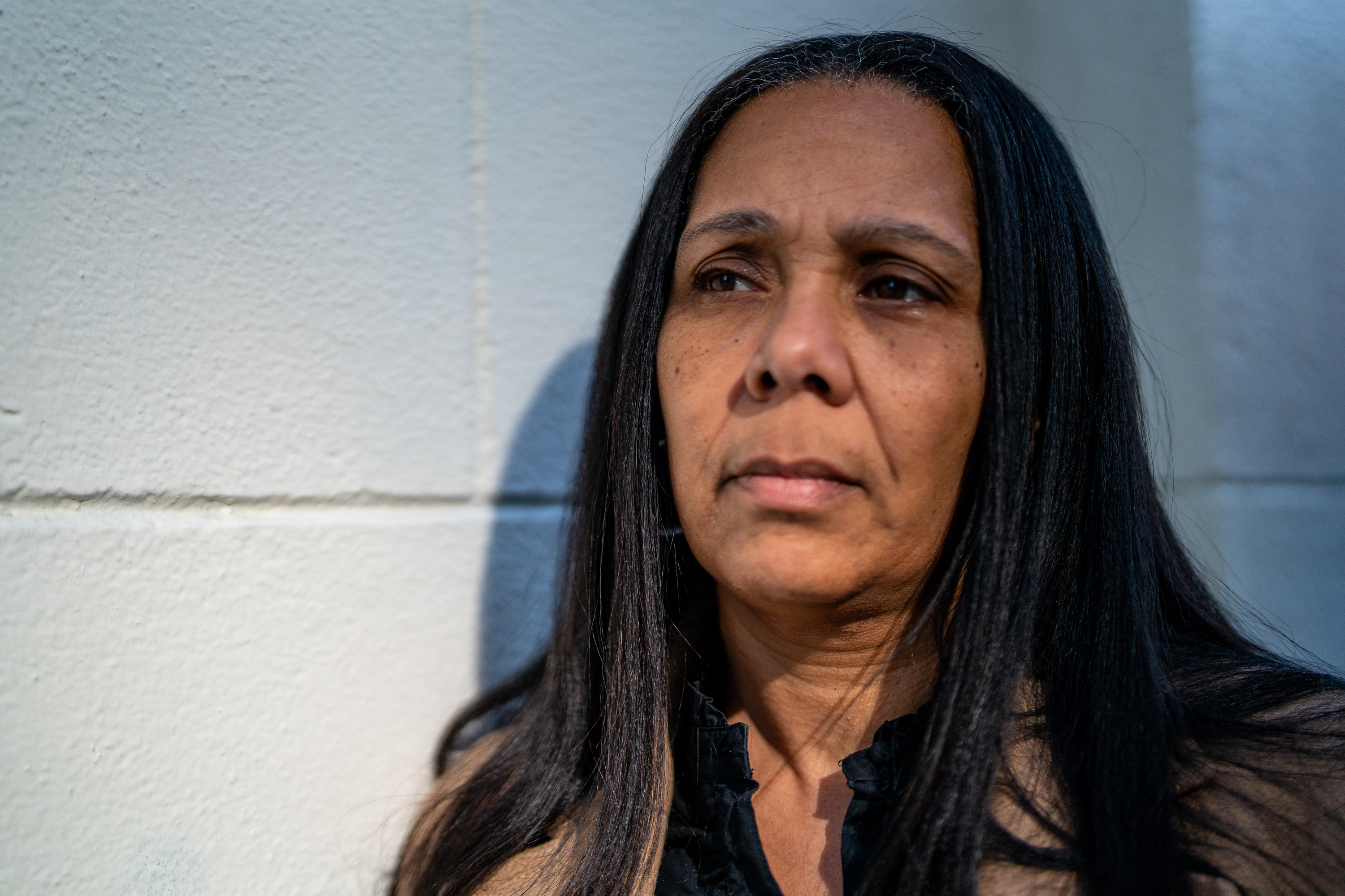 A woman with long dark hair stands against a textured white wall, looking away pensively, with soft sunlight illuminating her face.