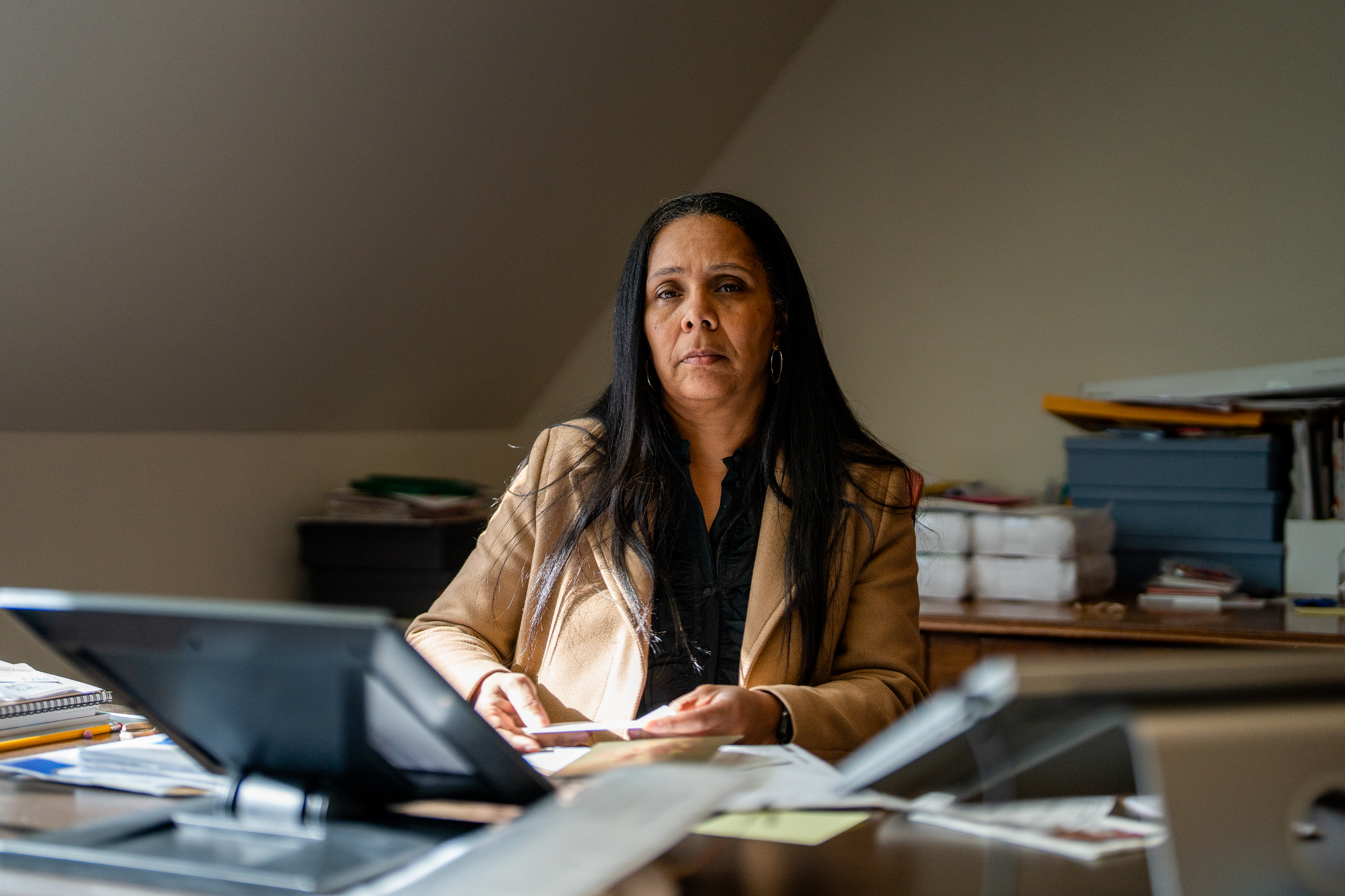 A woman with long dark hair sits at a desk in an office, wearing a tan blazer and looking towards the camera with a neutral expression.