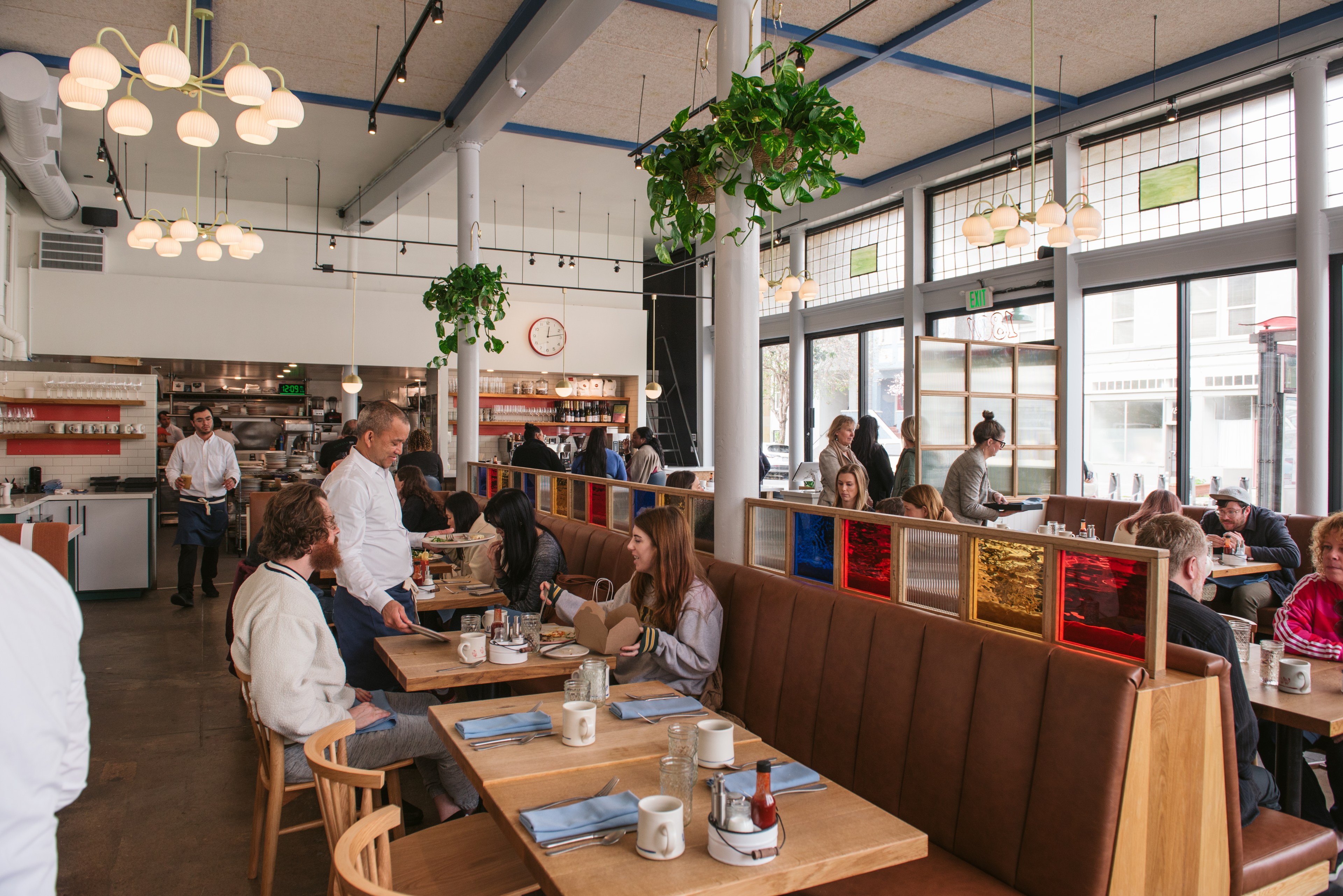 A bright, bustling café features wooden tables with blue napkins, people dining, hanging plants, and large windows. A server tends to customers while others chat.