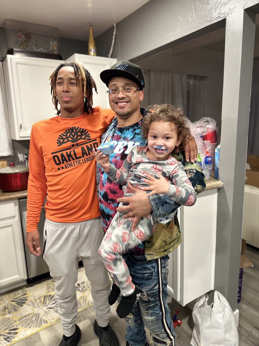 A joyful moment in a kitchen shows three people. A man in a colorful shirt holds a small child with blue icing on their face, next to a smiling person in an orange shirt.