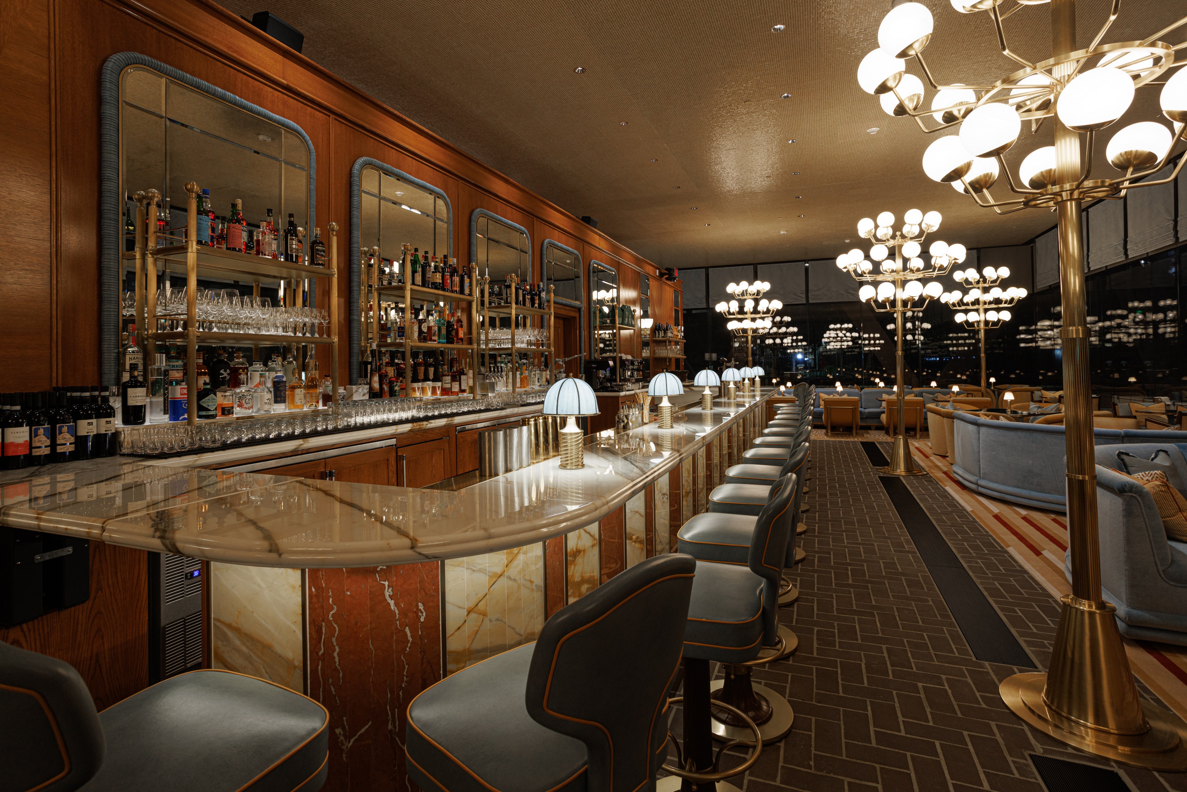 The image shows an elegant bar with a marble countertop and cushioned bar stools. Shelves behind hold various bottles and glasses, with ornate lighting overhead.