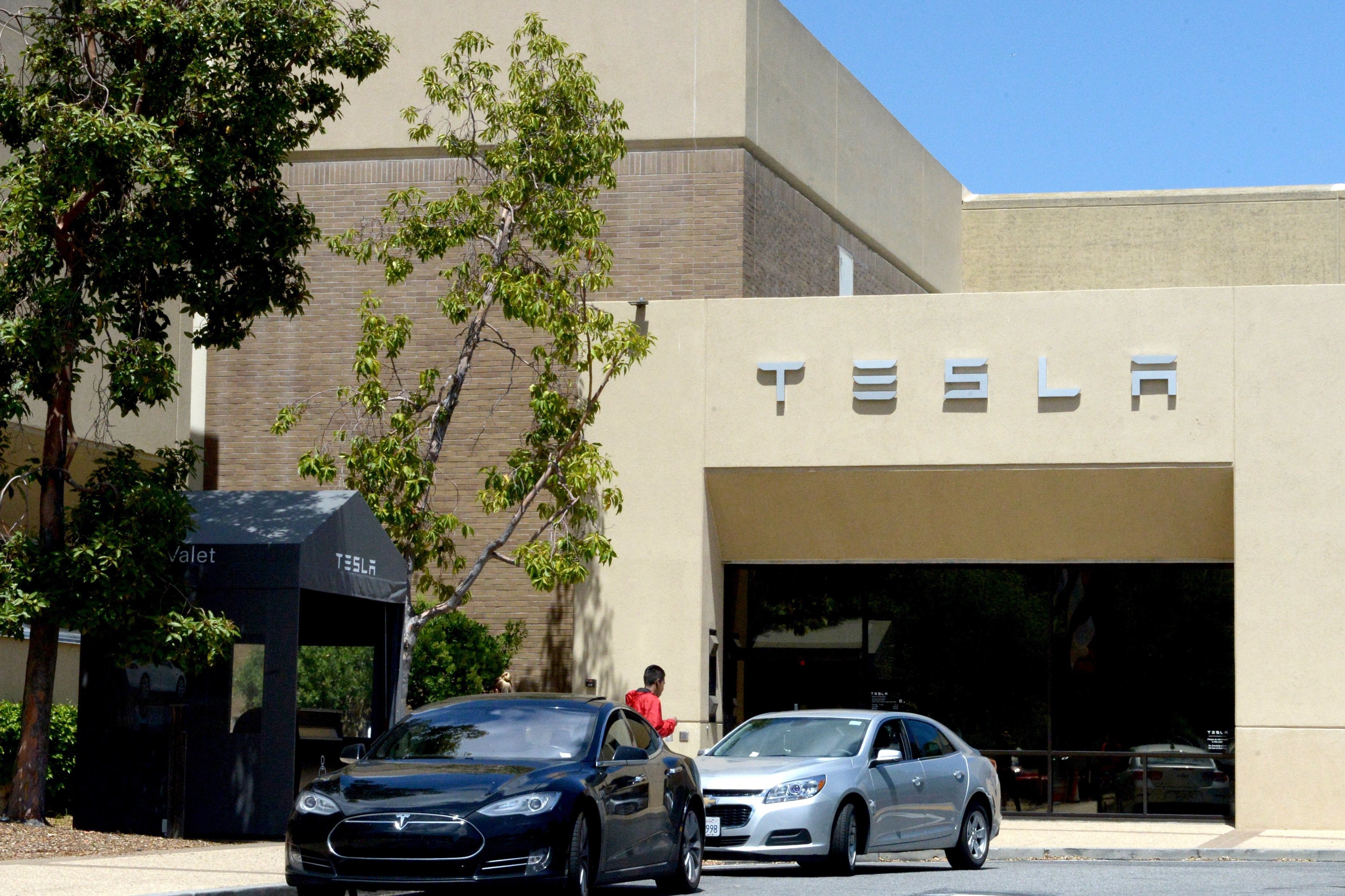 The image shows two cars, one black and one silver, parked near a building with the "TESLA" logo on it. There are trees and a valet booth nearby.