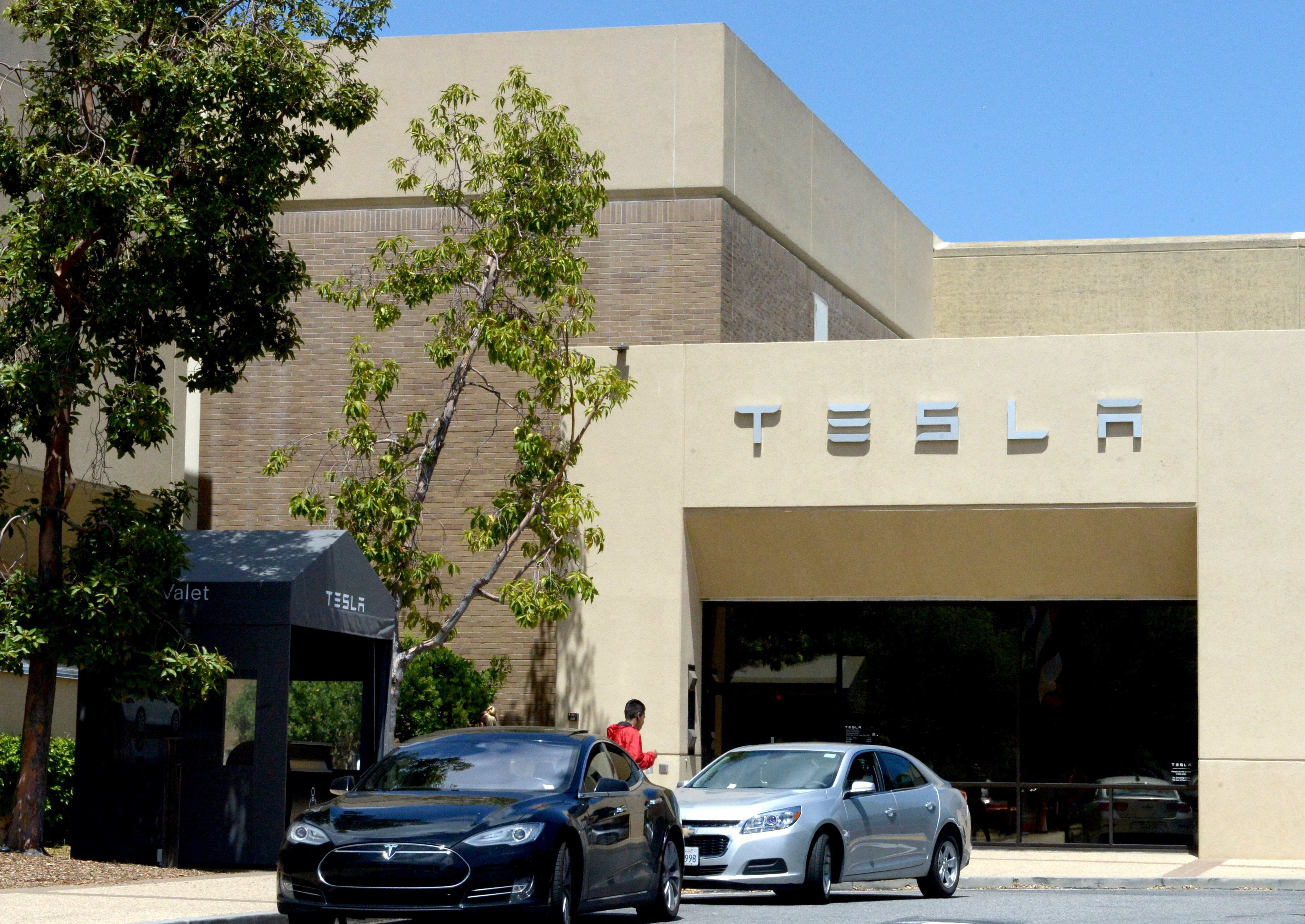 The image shows two cars, one black and one silver, parked near a building with the "TESLA" logo on it. There are trees and a valet booth nearby.