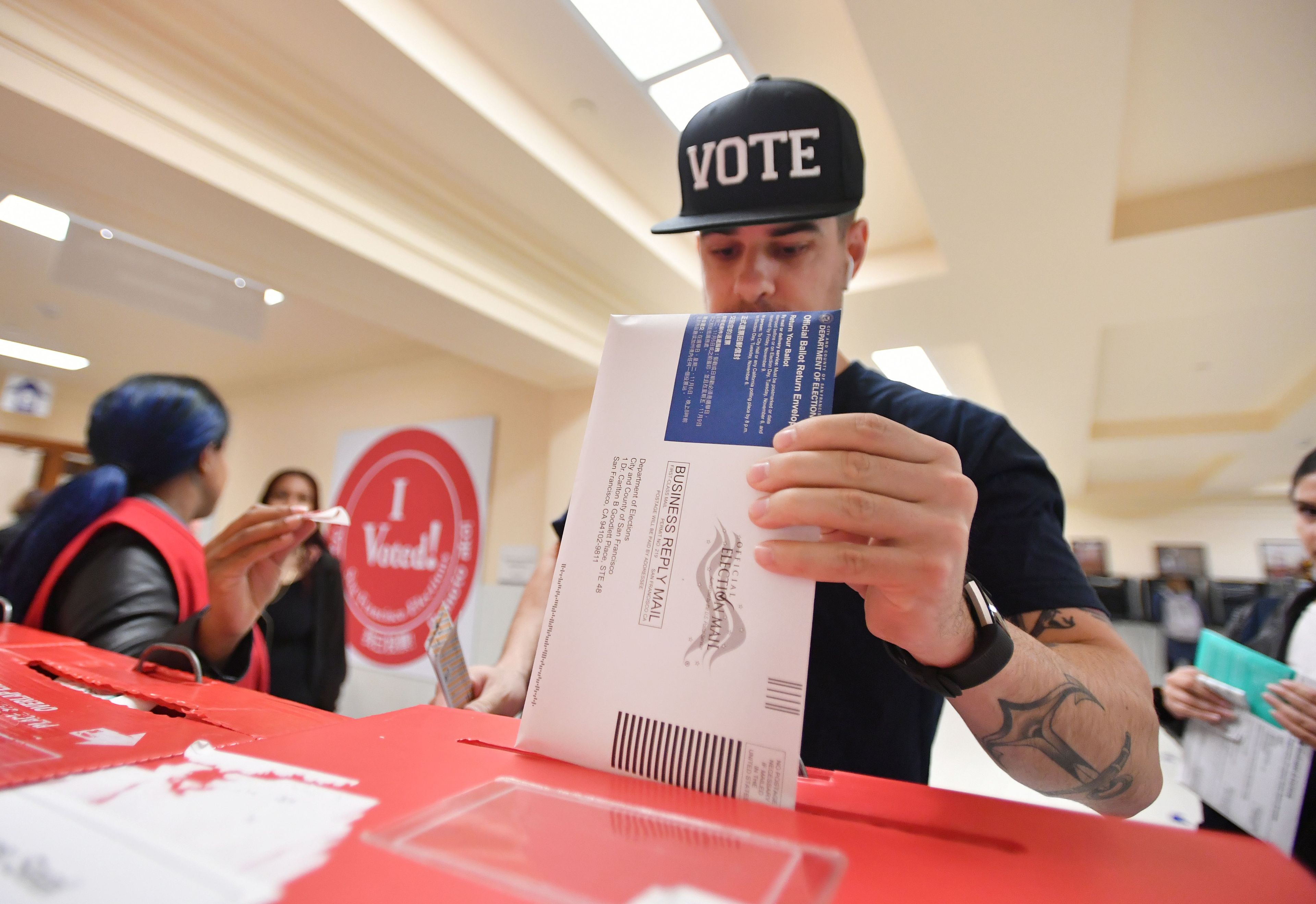 A man wearing a hat drops a ballot envelope into a container.