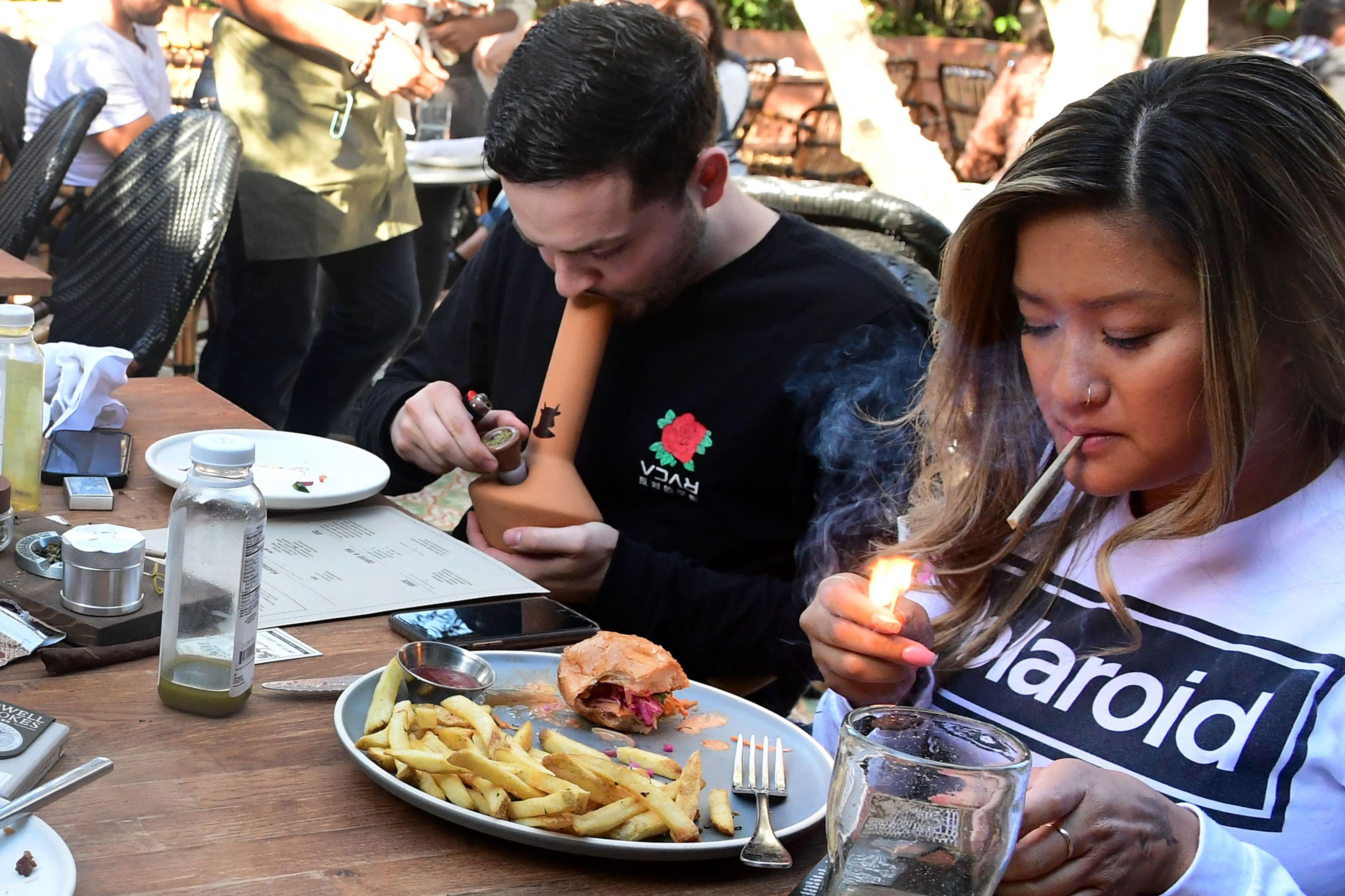 A woman lights a cigarette while a man uses a smoking device. They are seated at a table with food, including fries and a sandwich, and various drink bottles.