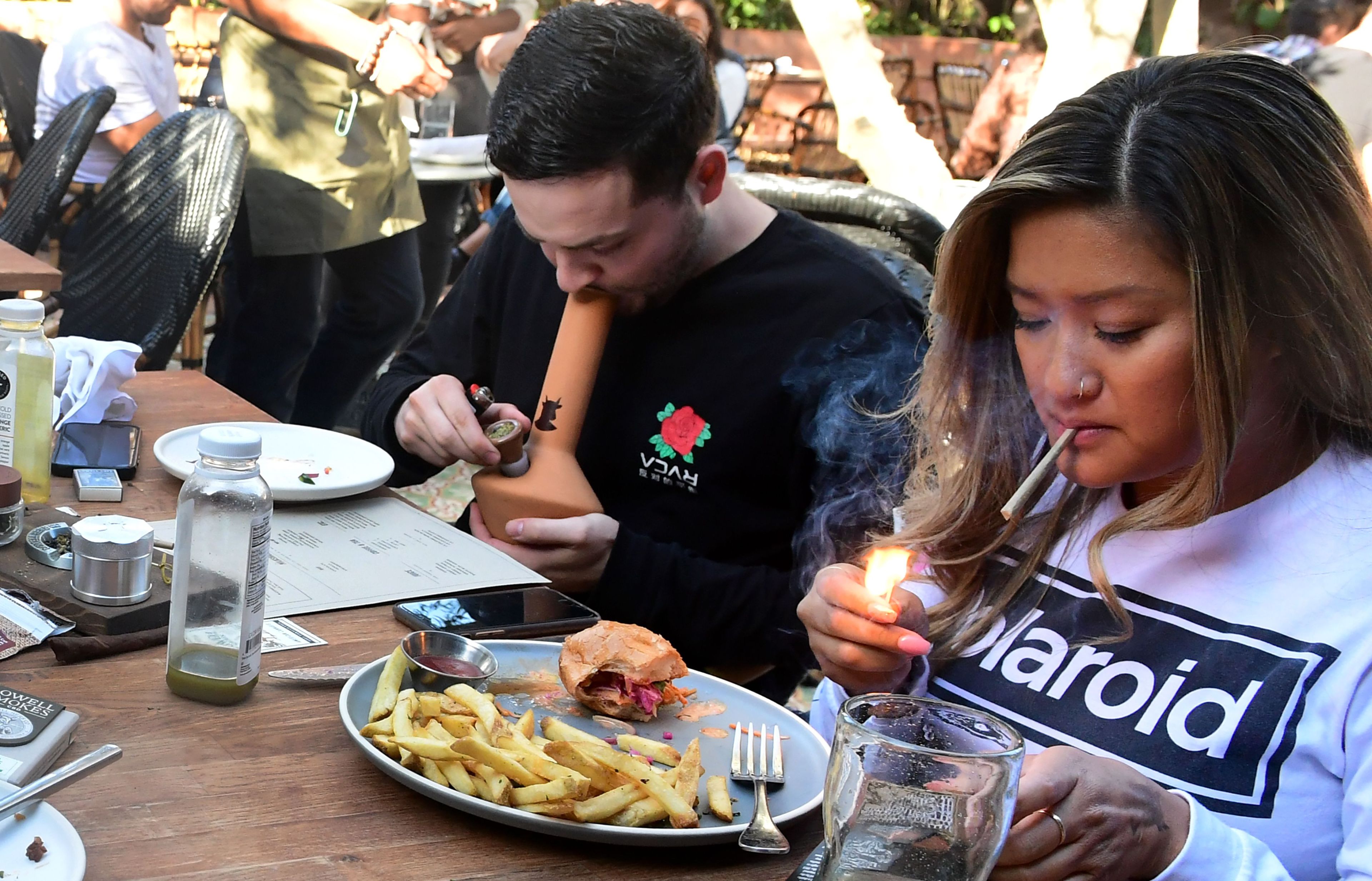 A woman lights a cigarette while a man uses a smoking device. They are seated at a table with food, including fries and a sandwich, and various drink bottles.