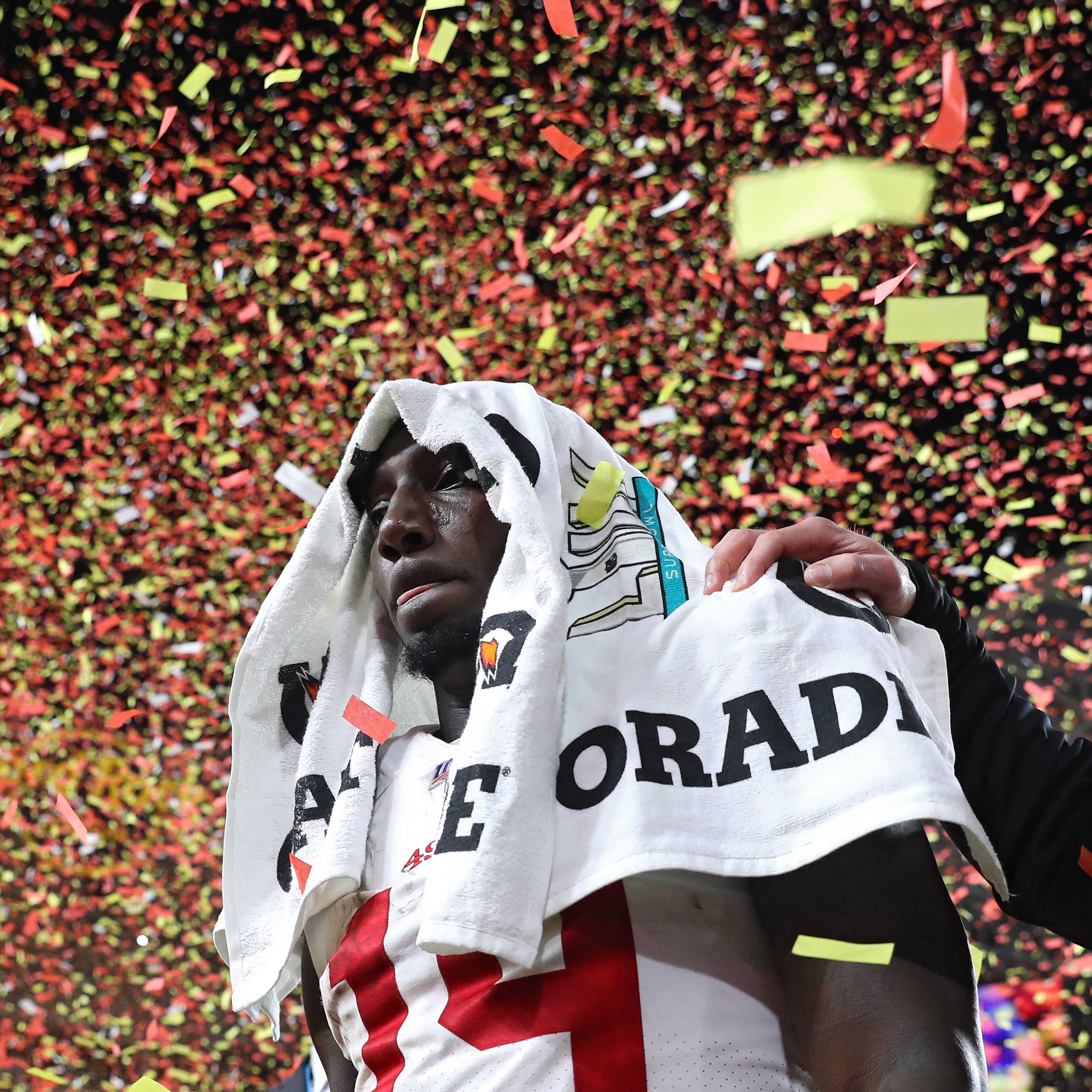 A football player stands with a towel over his head amidst falling red and yellow confetti. A supportive hand rests on his shoulder, suggesting a celebratory moment.