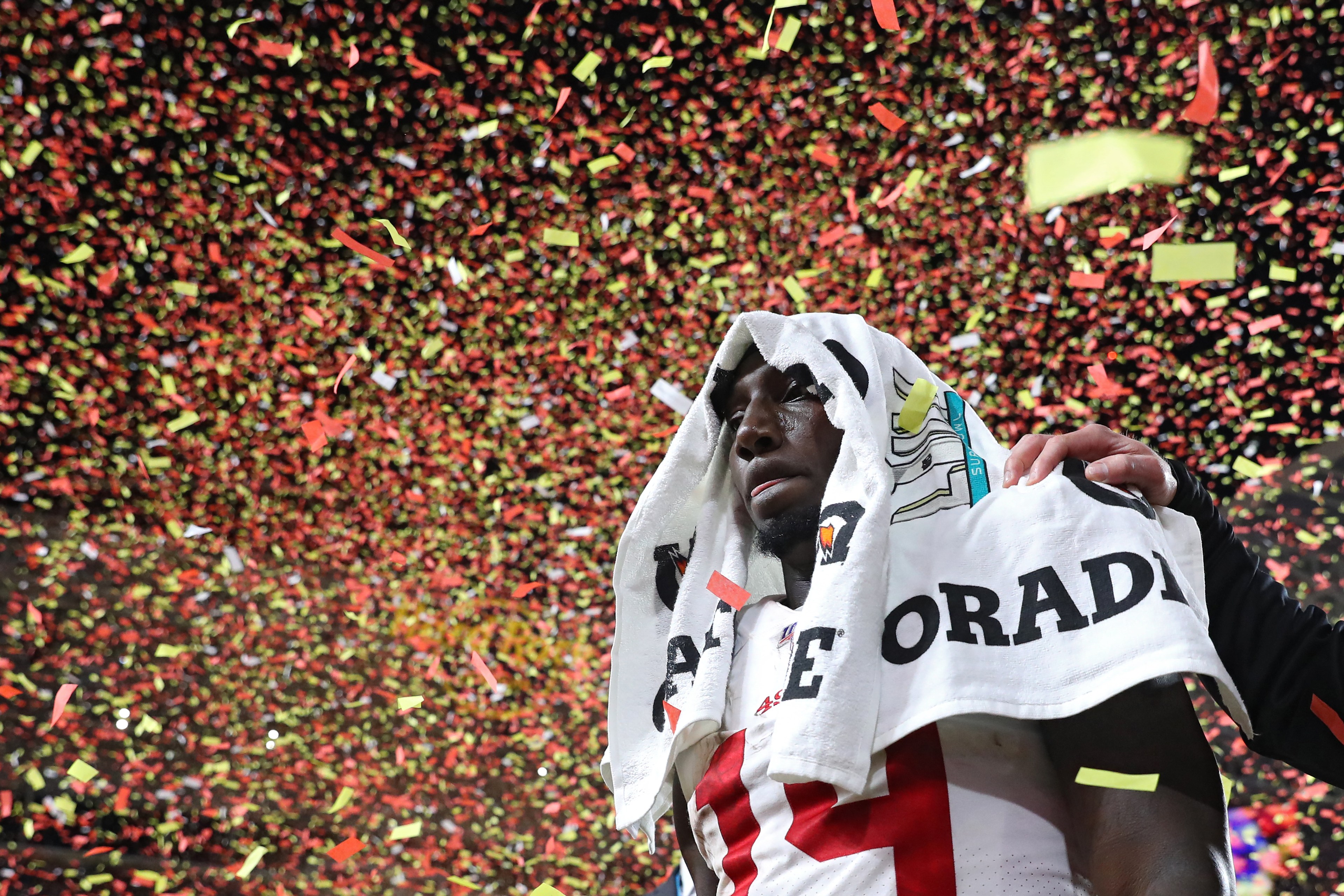 A football player stands with a towel over his head amidst falling red and yellow confetti. A supportive hand rests on his shoulder, suggesting a celebratory moment.