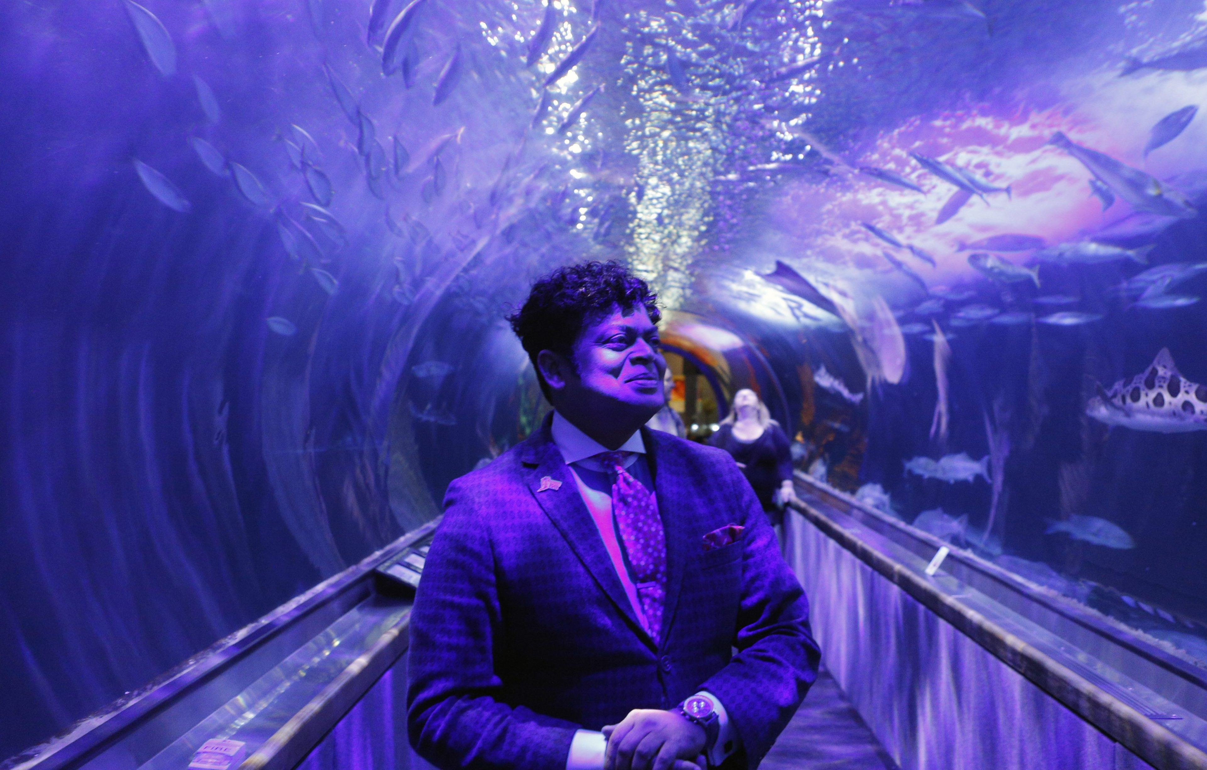 A man in a suit stands in an illuminated aquarium tunnel surrounded by swimming fish and a shark. The scene is bathed in purple and blue lighting.