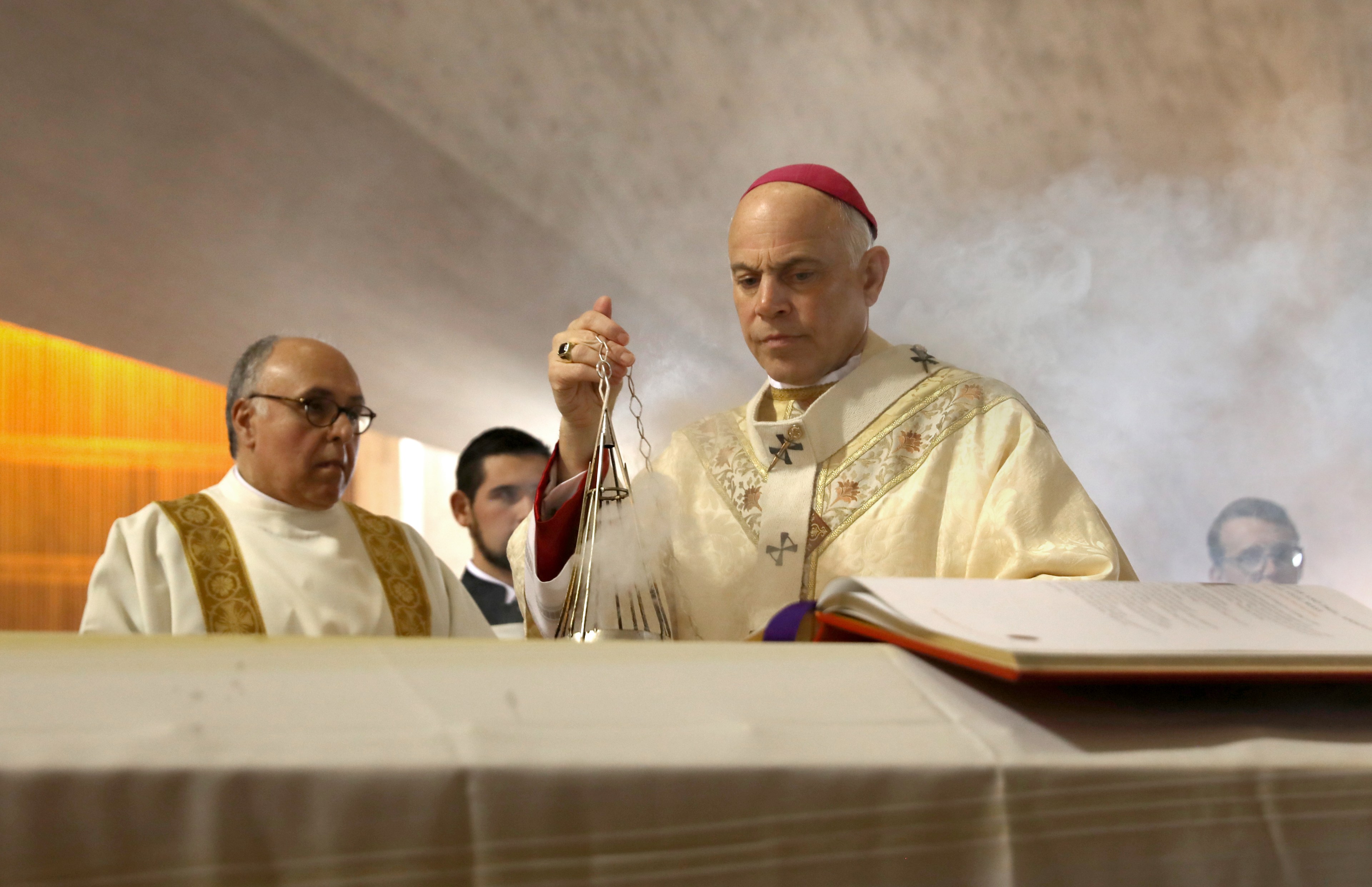 A religious ceremony scene with a clergyman in ornate robes holding a censer, releasing smoke. An altar with an open book is in the foreground.